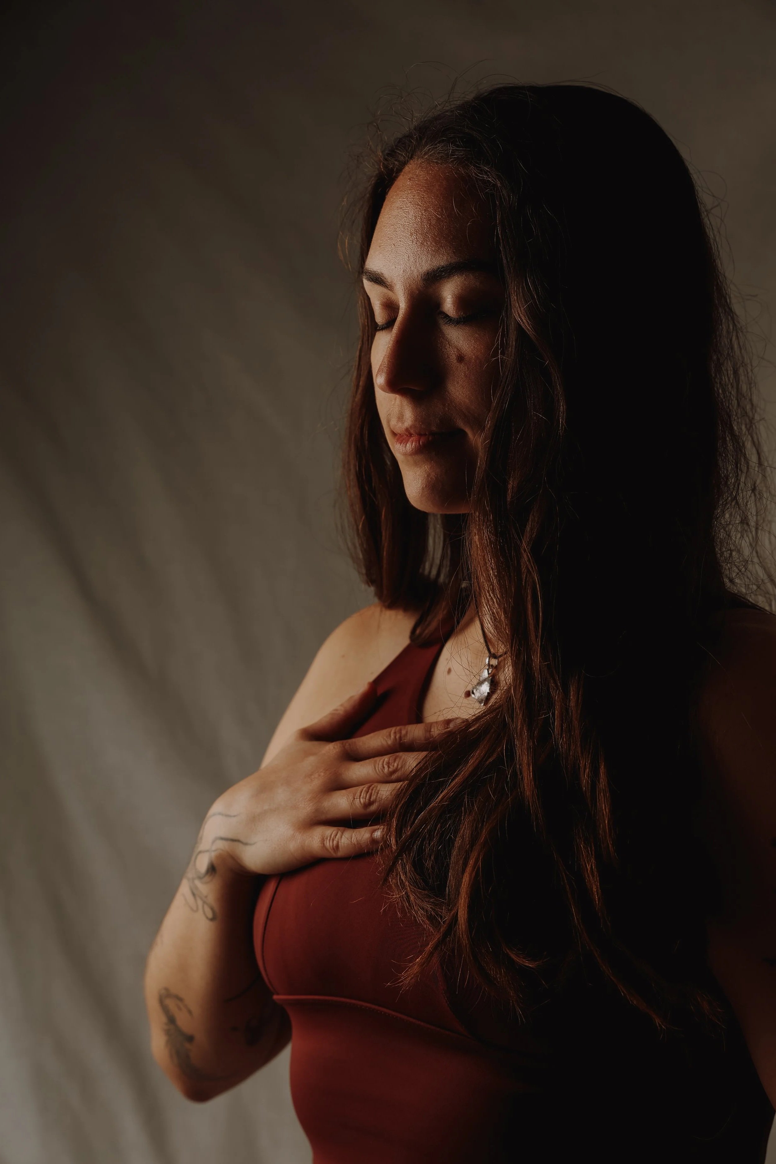 Eine junge Frau mit langem braunem Haar trägt ein rotes Top und hält ihre rechte Hand auf ihre Brust, während sie mit geschlossenen Augen nachdenklich wirkt.