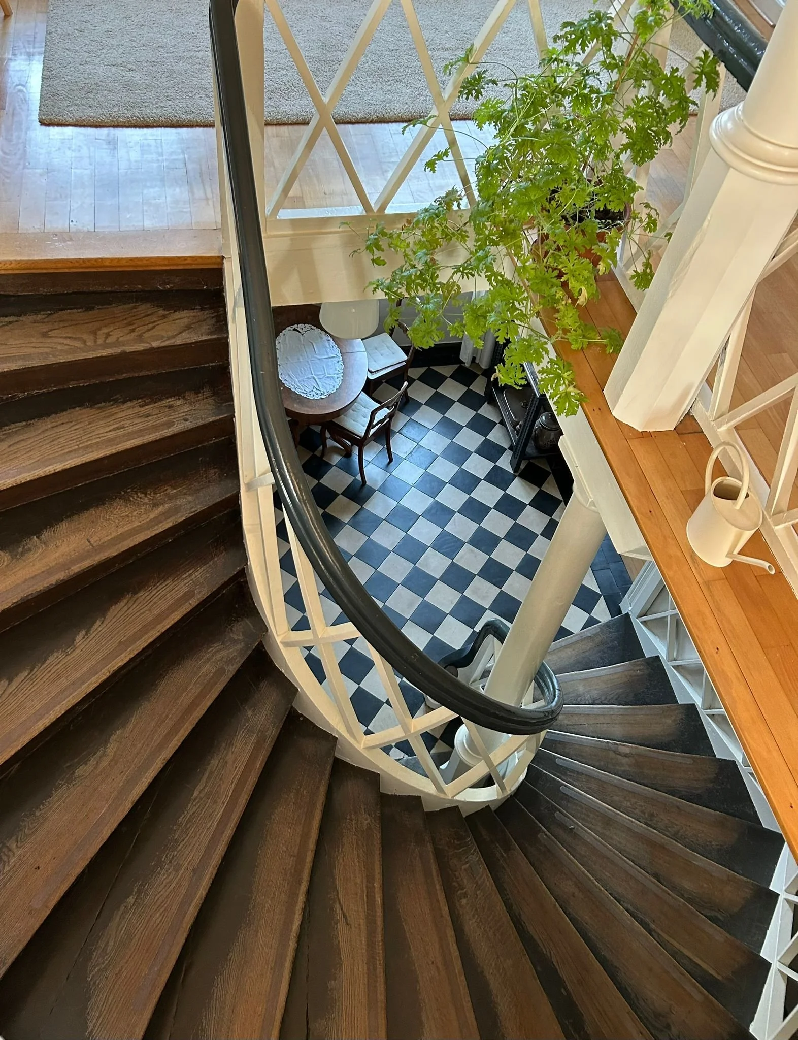 Blick nach unten auf eine Holztreppe einer viktorianischen Villa mit dunklen Holzbodenstufen, weiße Geländer und ein Dschungel von grünen Pflanzen oben. Unten ist ein Essbereich mit schwarzen und weißen Fliesen, einem kleinen runden Tisch, Stühlen un