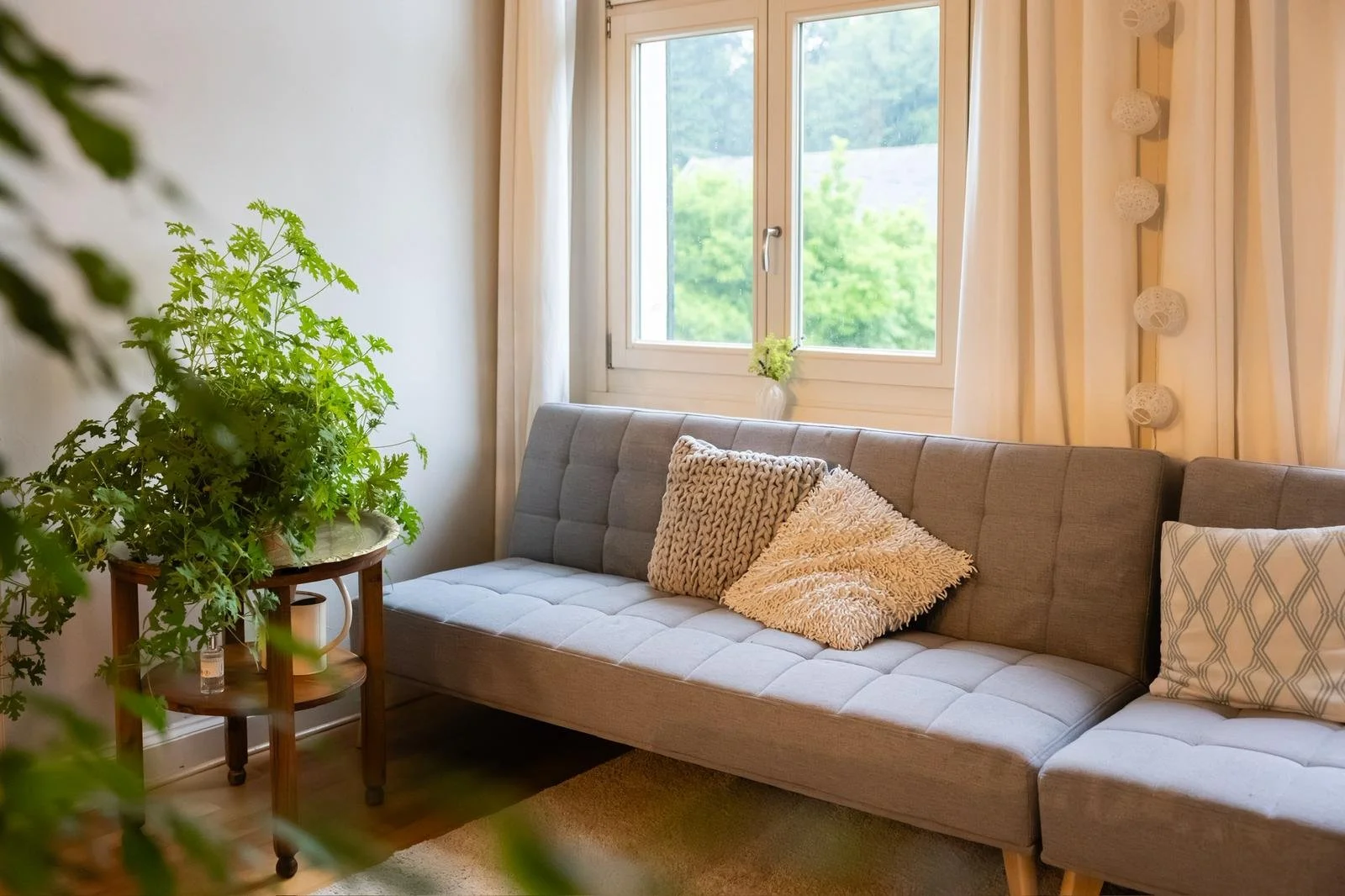 Ein gemütliches Wohnzimmer mit einem grauen Sofa, Kissen und einer Fensterfront, durch die grüner Ausblick ins Freie sichtbar ist.