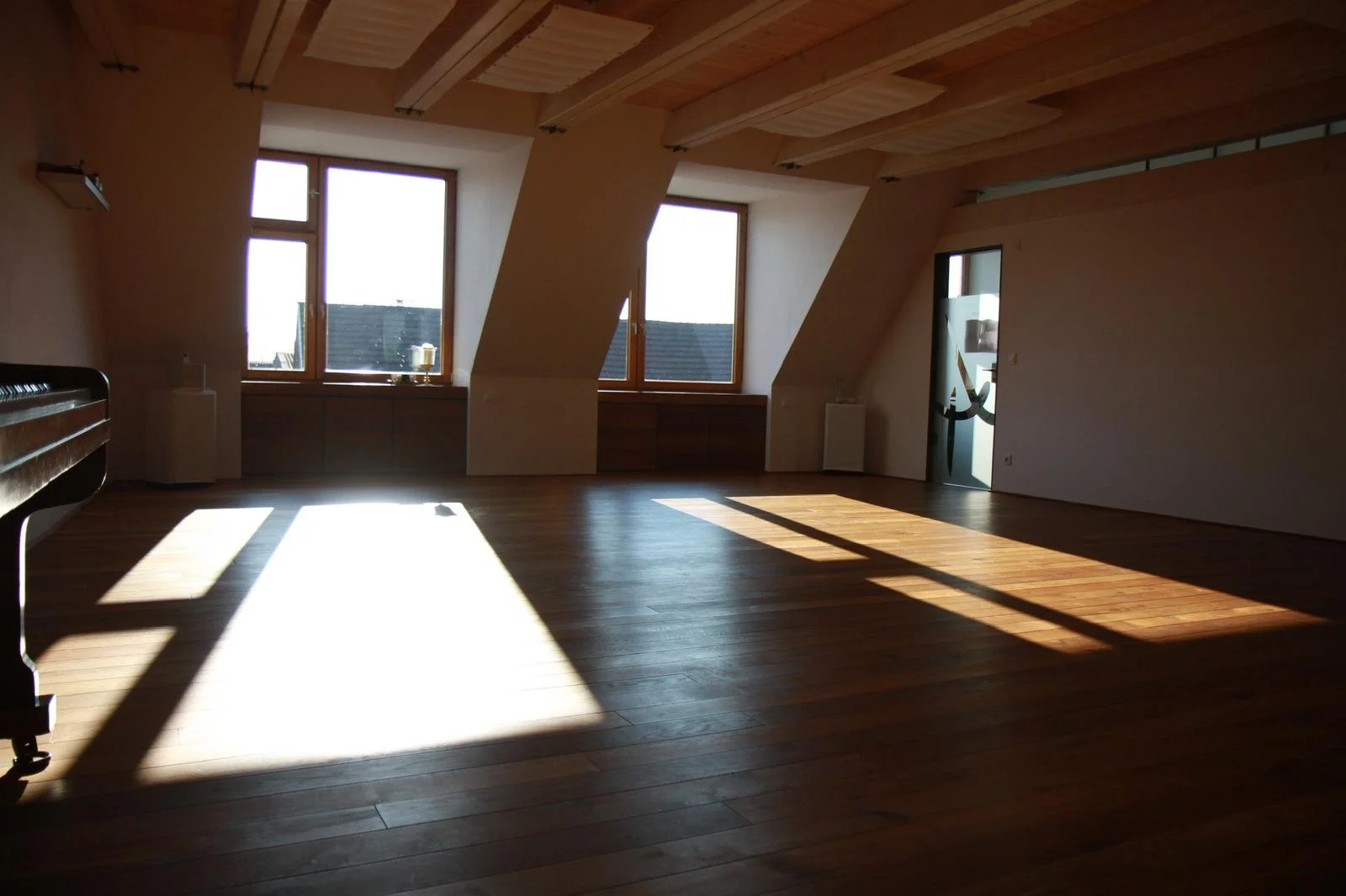 Ein leeres Dachgeschosszimmer mit Holzboden und großen Fenstern, durch die Sonnenlicht auf den Boden fällt. An der linken Wand steht ein Klavier.