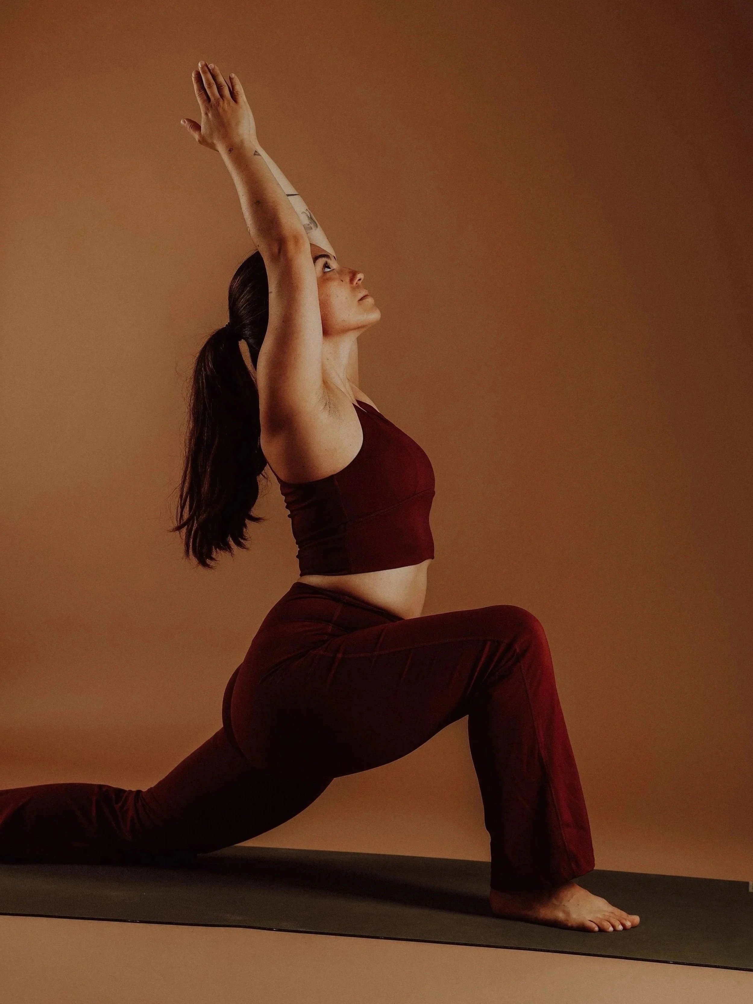 Frau praktiziert Yoga in der Zugstellung auf einer Yogamatte, trägt dunkle Sportkleidung, dunkle Haare, Blick nach oben gegen die Wand.