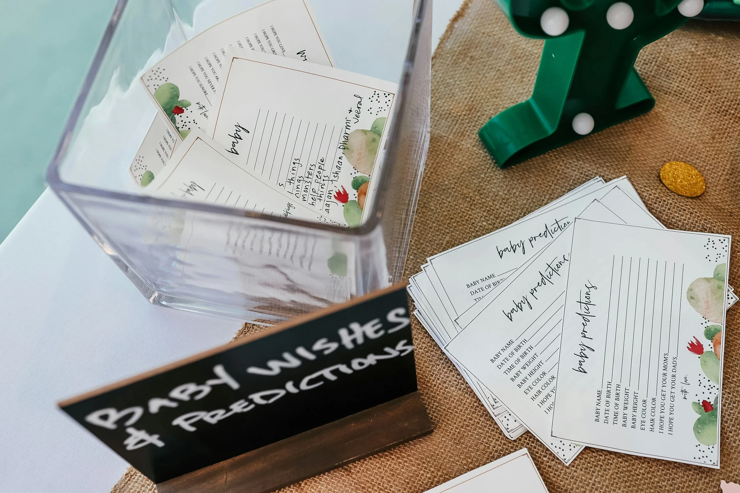 Karten für Baby Vorhersagen in einer Glasbox, auf einem braunen Tuch, mit einem grünen Stempel und einer goldenen Scheibe daneben.
