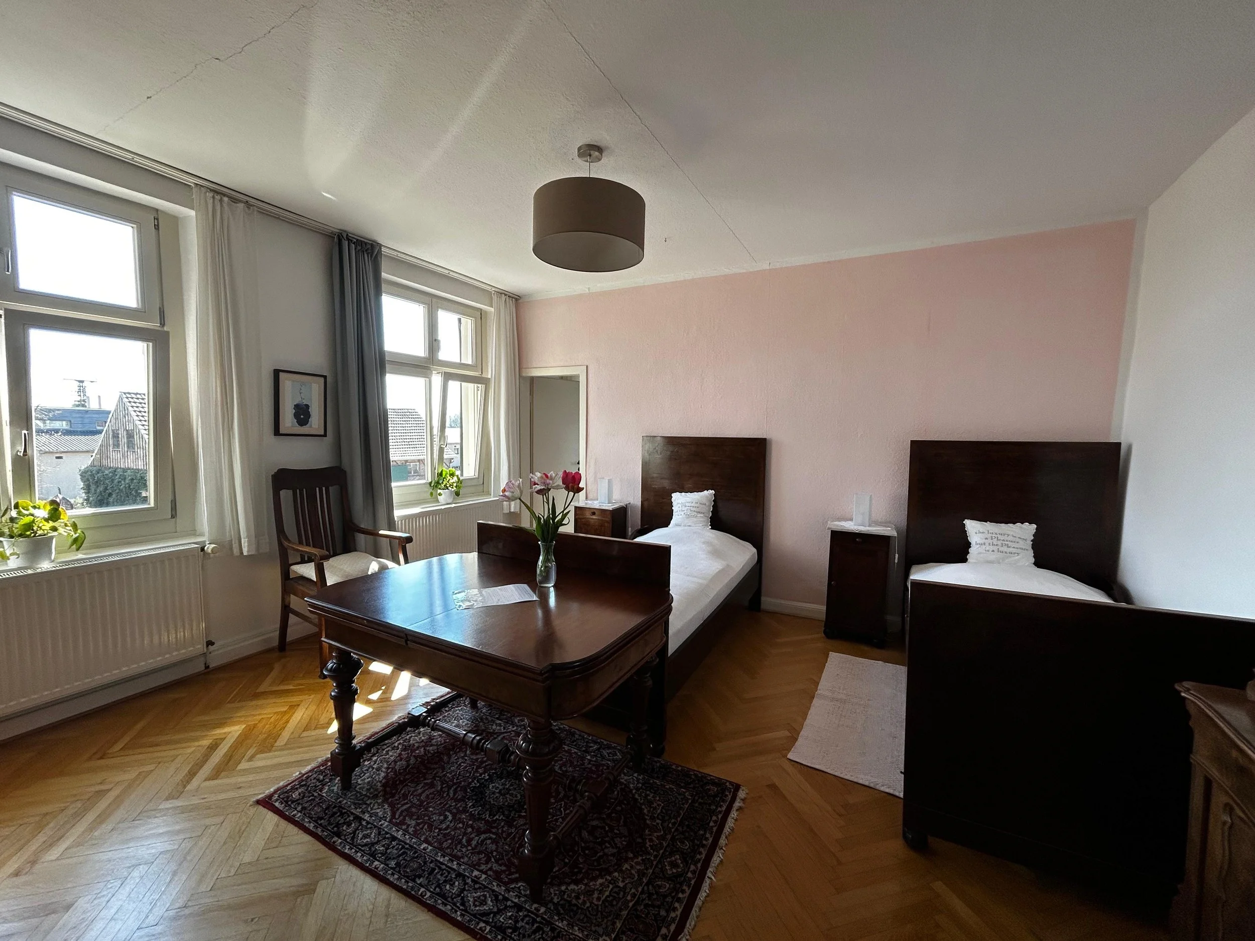Zimmer mit zwei Einzelbetten, einem großen Fenster mit weißen und grauen Vorhängen, einem Holz Tisch mit Vase und Tulpen, einem Sessel, einem Teppich auf Holzboden, Wände in Weiß und Hellrosa, und einem Deckenlampenschirm.