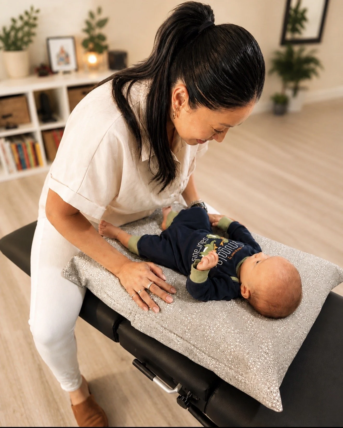 A woman is leaning over a baby lying on a changing table, smiling and looking at the baby. The baby is dressed in a dark blue outfit and is looking up. The room has a bookshelf with books and plants in the background.
