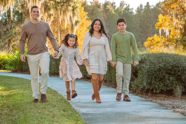 A family of four walking hand-in-hand outdoors on a park path during fall, with trees and bushes in the background.