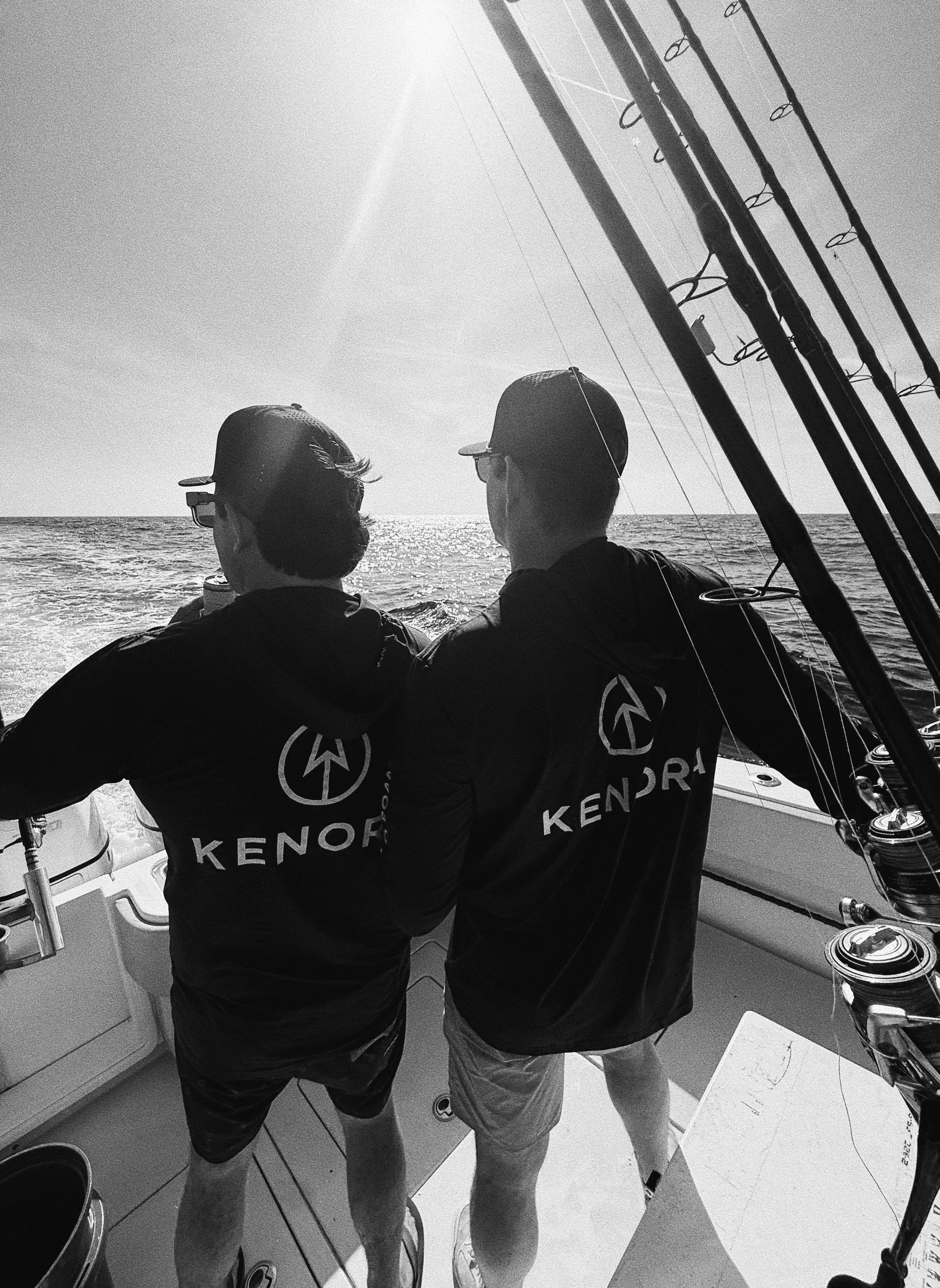 Two men wearing hoodies with 'KENDOR' on the back, standing on a boat, watching the sunset over the ocean.