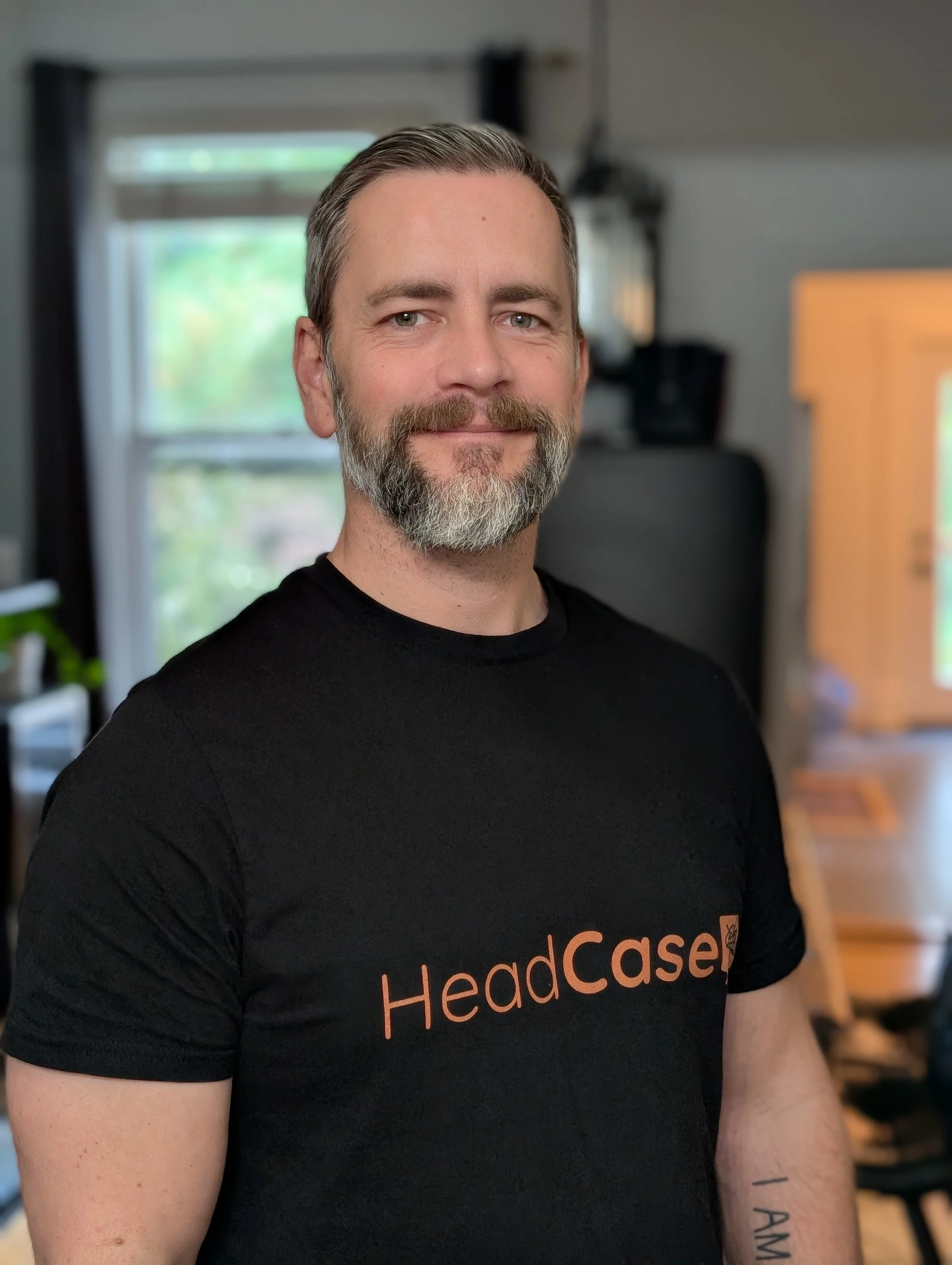 A man with short hair, a beard, and a mustache, smiling, wearing a black t-shirt with 'HeadCase' written on it in orange, standing indoors with a window and some furniture in the background.