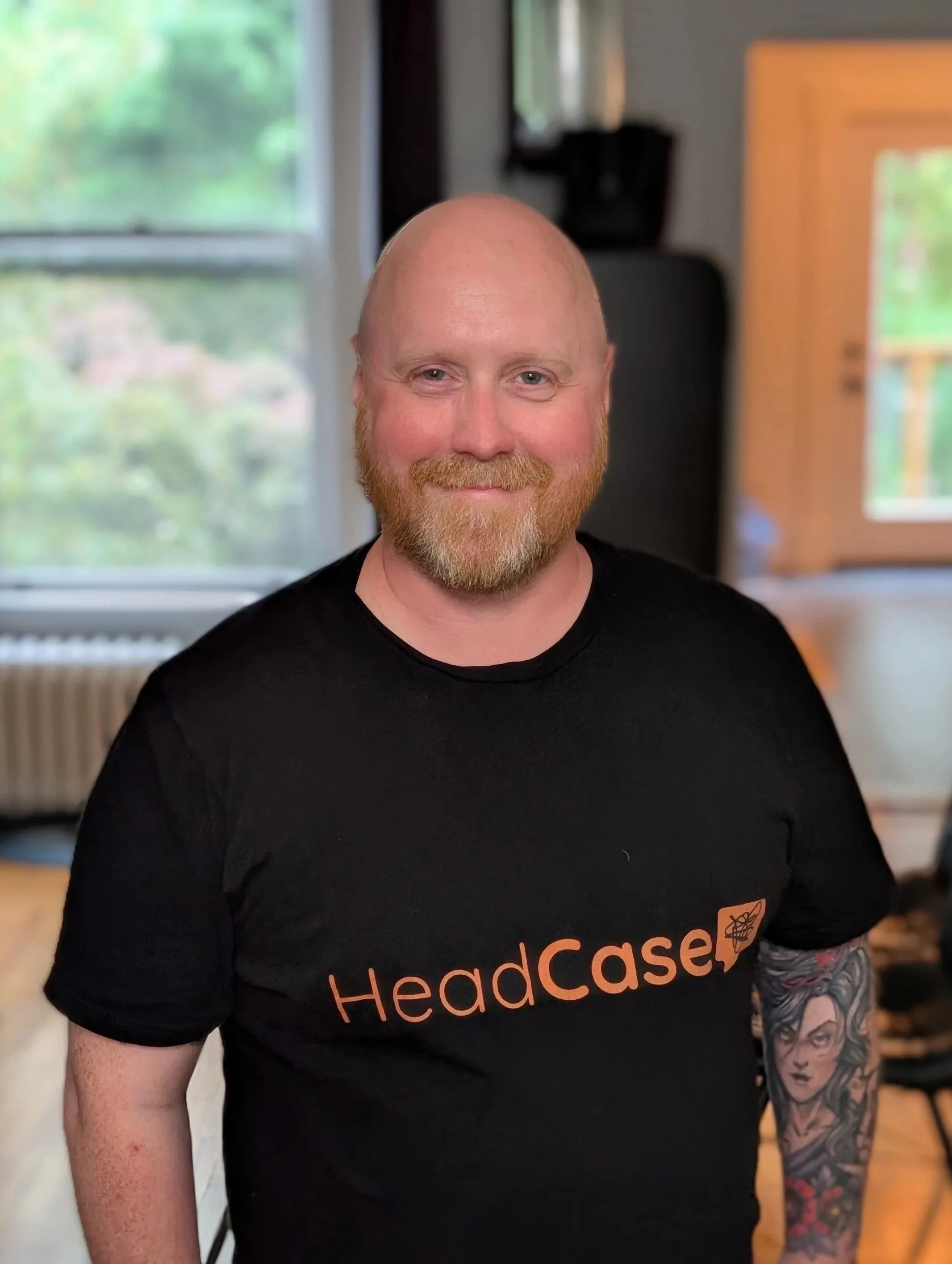 A smiling man with a beard and tattoos wearing a black t-shirt with the words 'HeadCase' during daytime indoors.