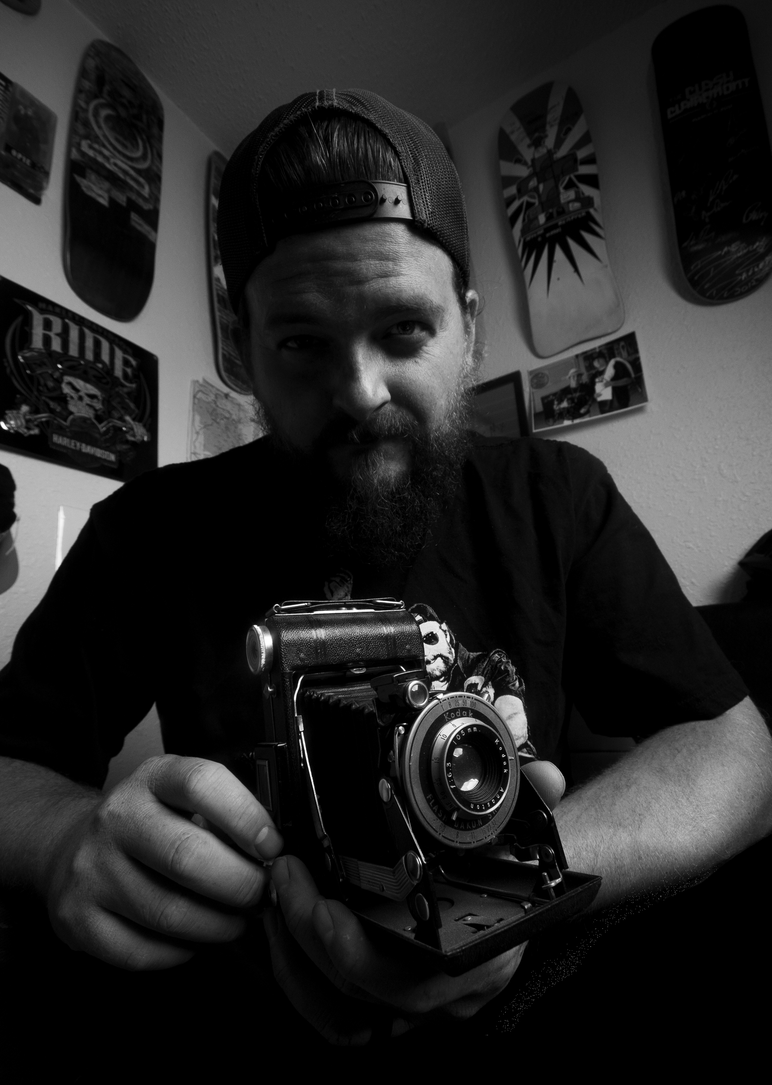 A man with a beard and a cap backwards, holding an old-fashioned camera, sitting indoors with skateboard art on the wall behind him.