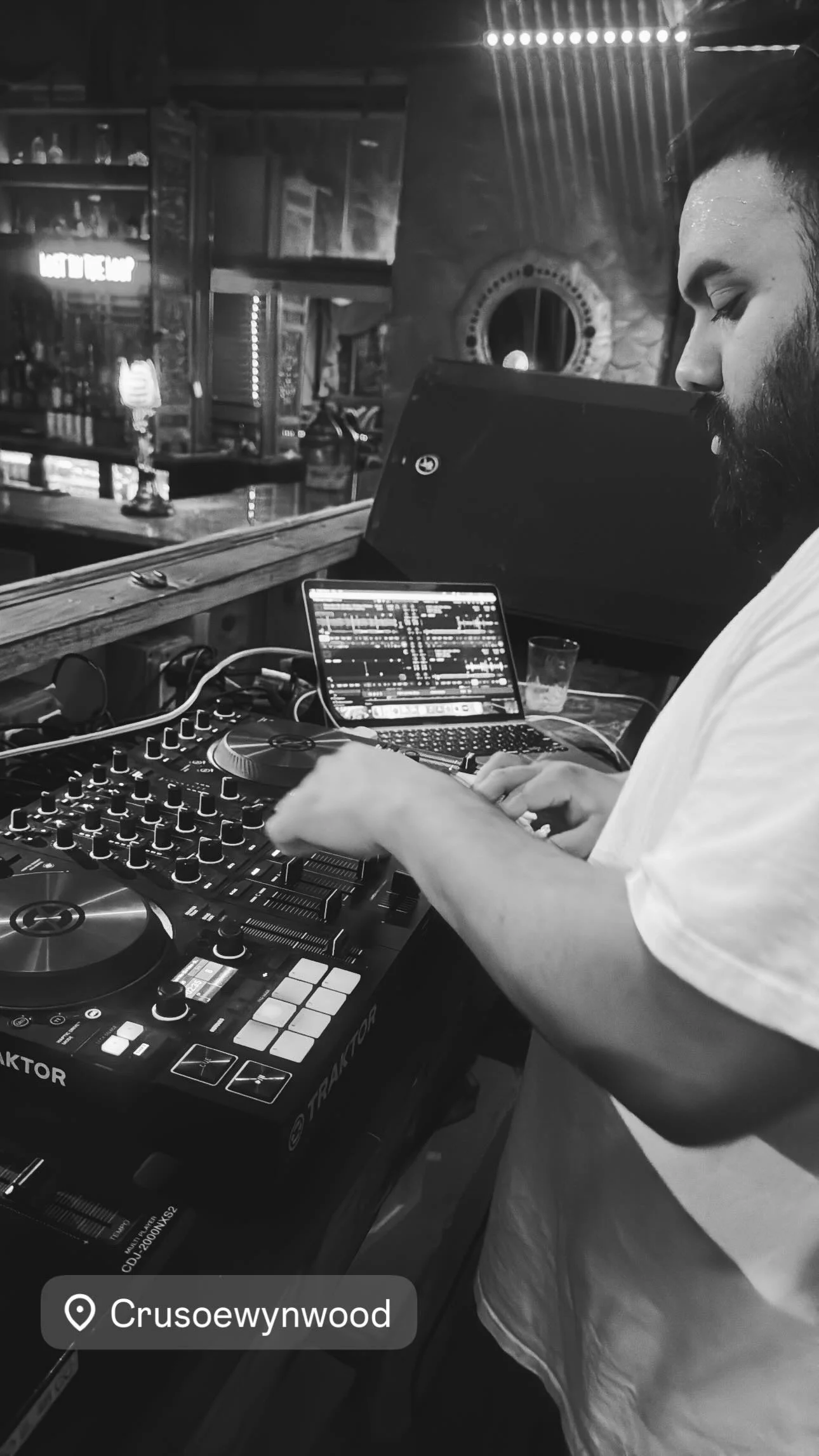A DJ with a beard mixing music on a DJ controller at a bar or club with a laptop displaying music software, in a dimly lit environment