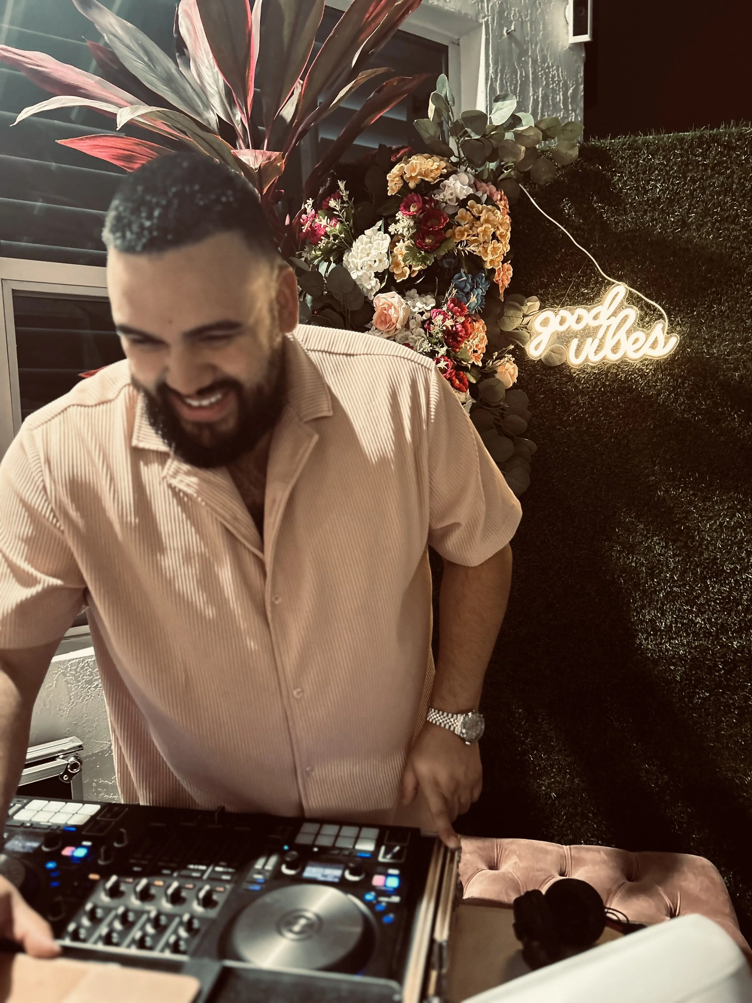A man with a beard and short dark hair smiling while operating a DJ controller at a party or event. Behind him are colorful flowers, green leaves, a window, and a neon sign that reads 'good vibes'.