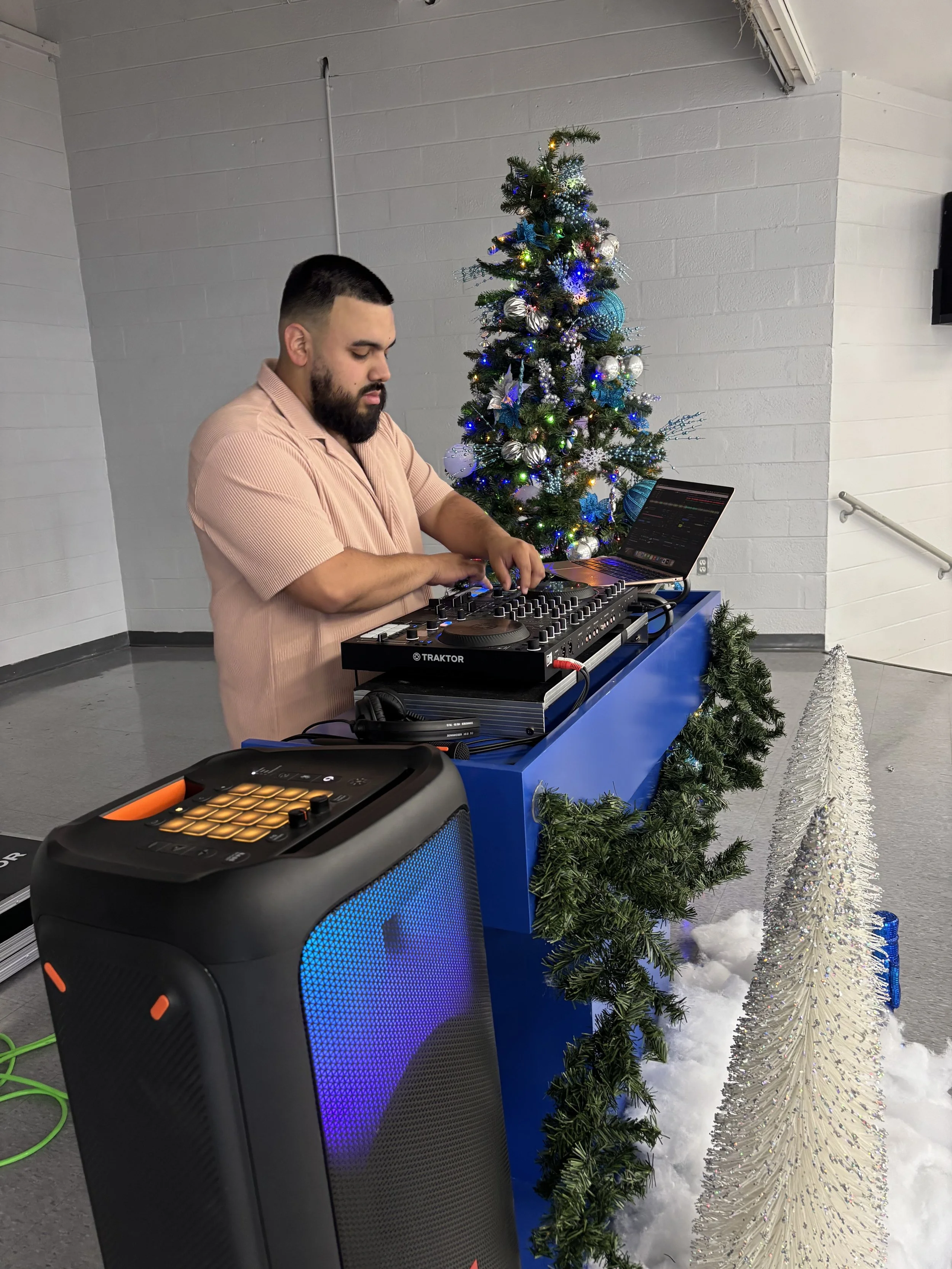 A man with a beard and short dark hair wearing a pink short-sleeved shirt DJing at a holiday party, with a decorated Christmas tree, white tinsel tree, and snowy decor in the background.
