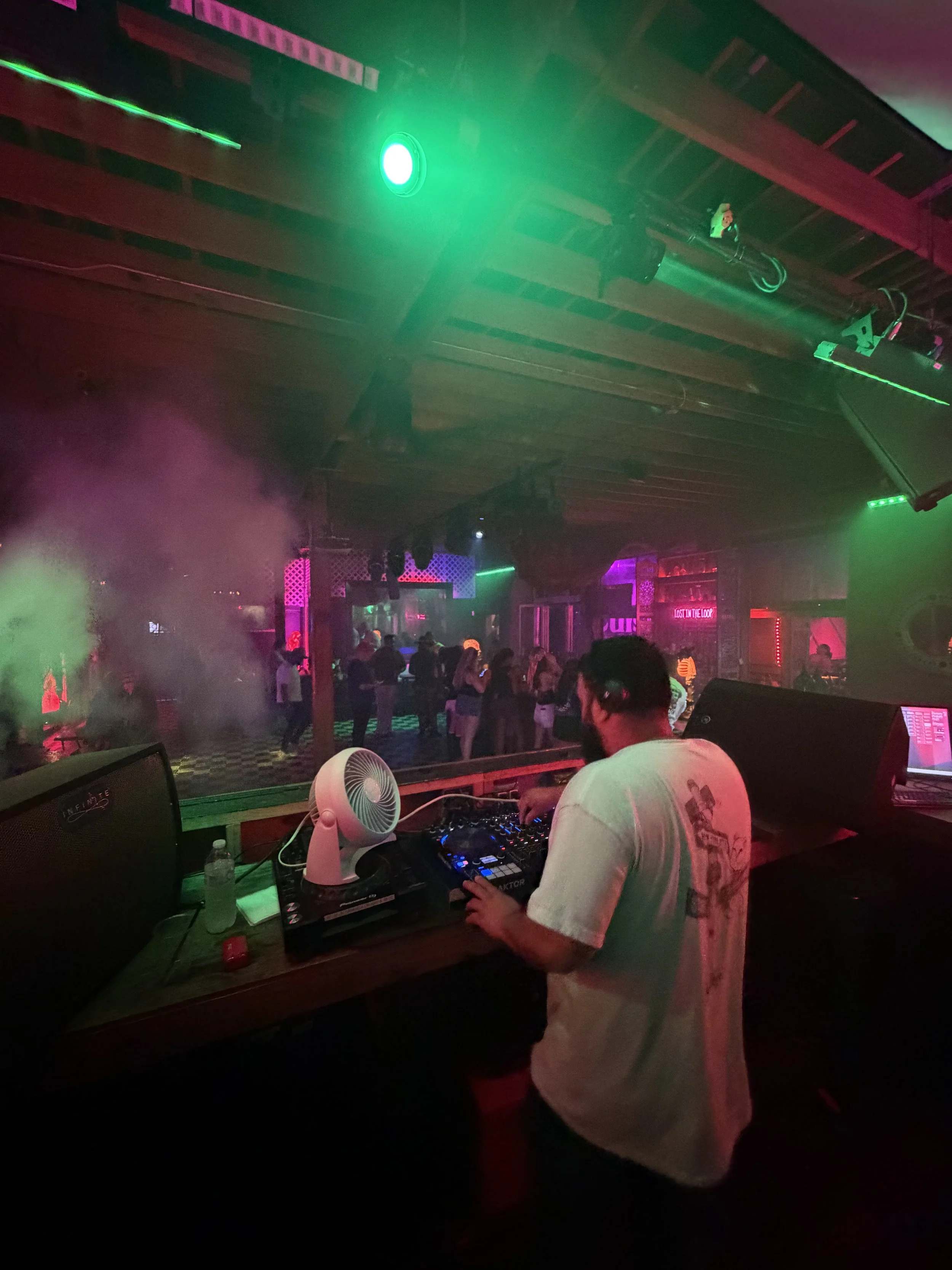 DJ performing at a nightclub with colorful lights and a dance floor with people dancing in the background.