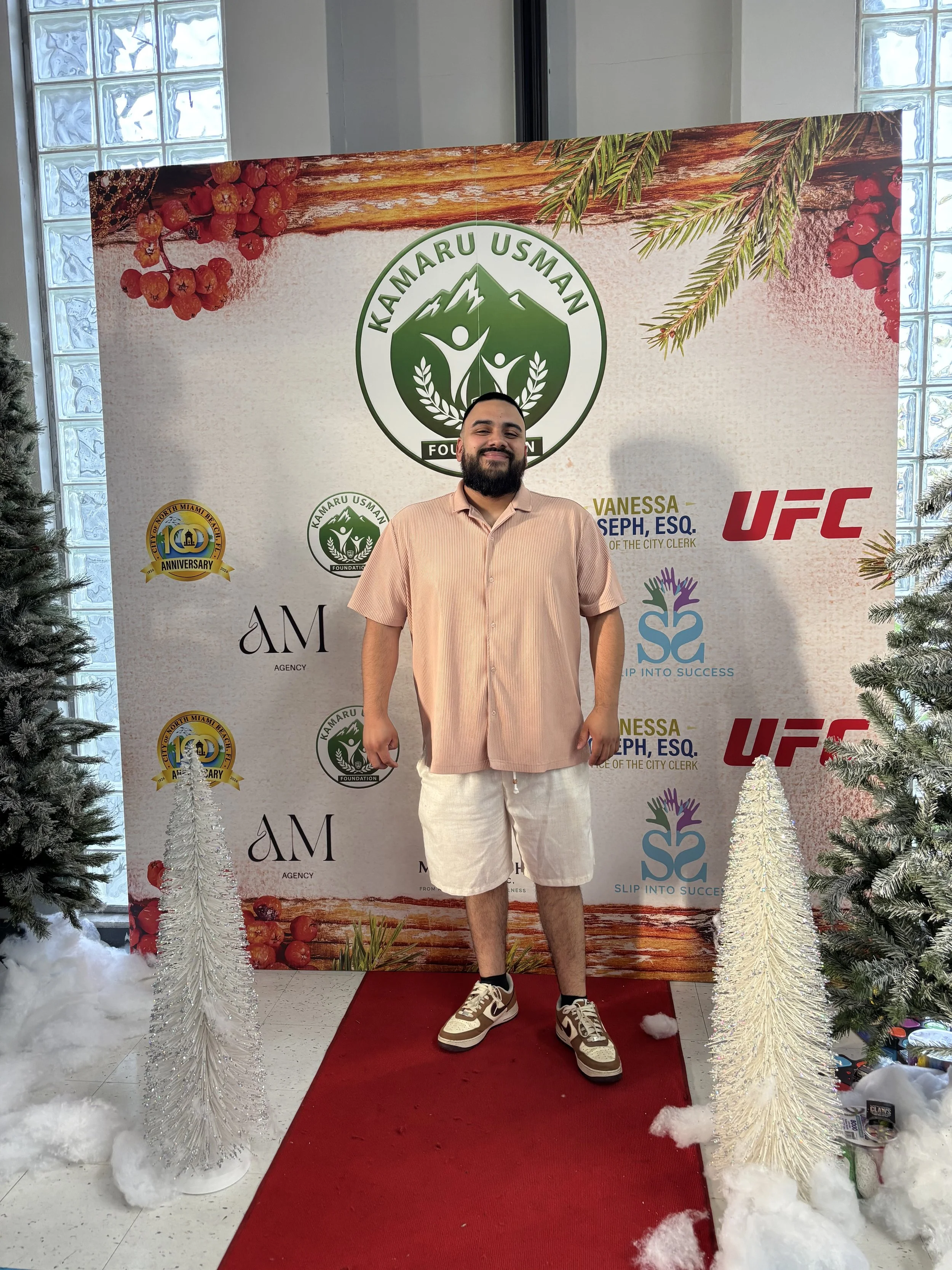 A man standing on a red carpet in front of a backdrop with logos, holiday decorations, artificial snow, and fake Christmas trees.