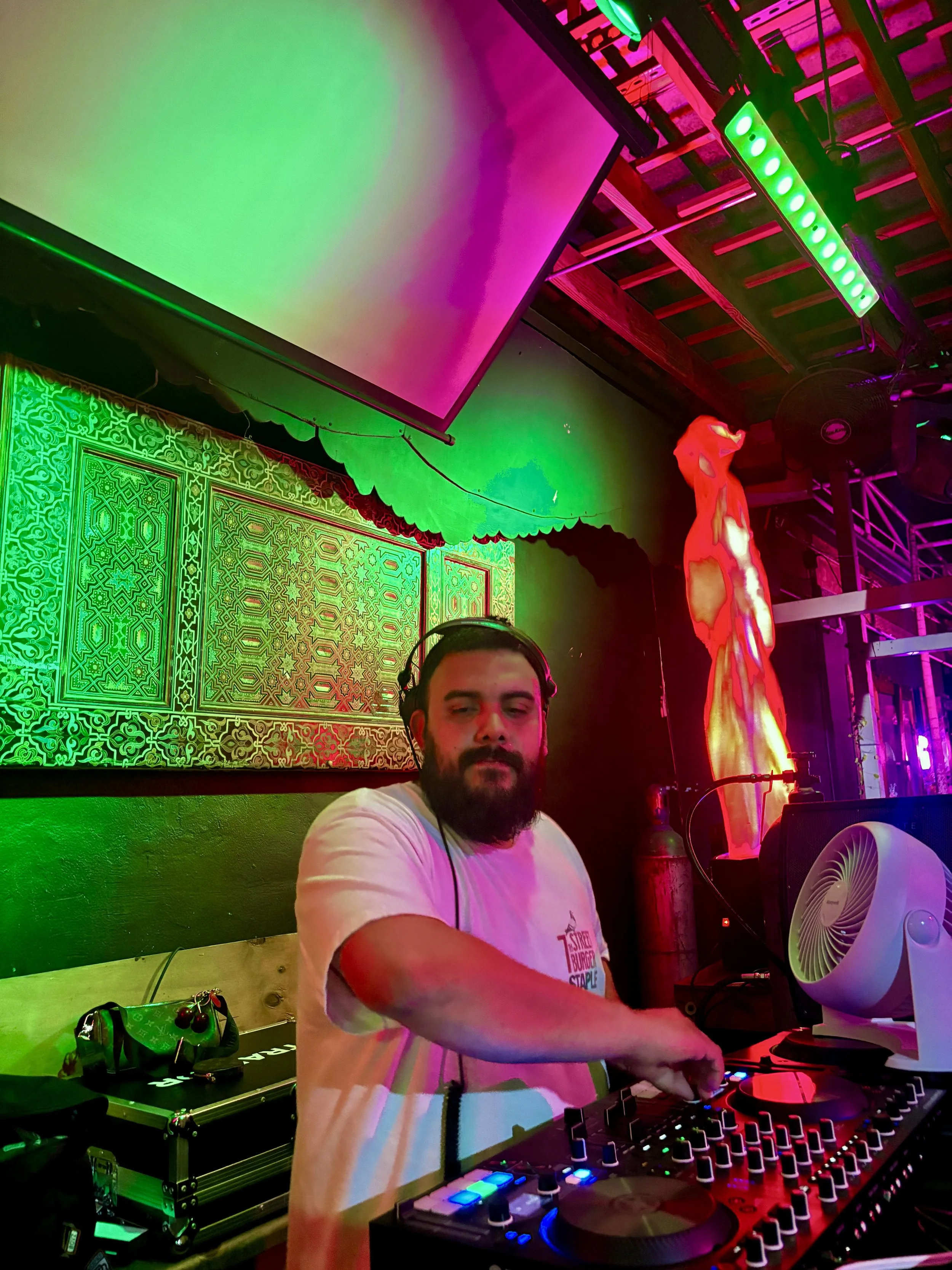 A DJ with a beard and headphones mixing music at a colorful club or event space. The background includes a patterned tapestry, a bright red statue, colored lights, and a large screen.
