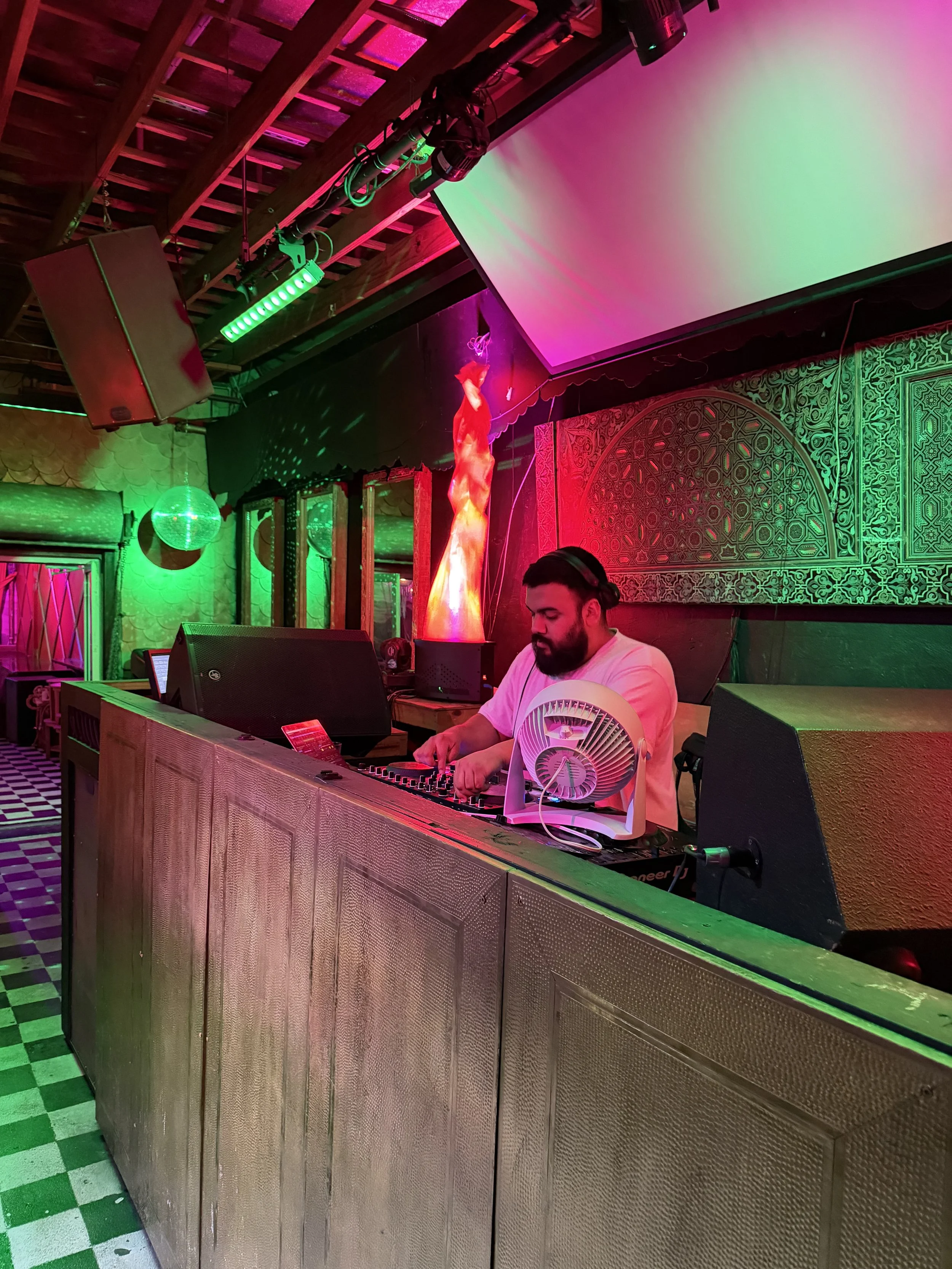 A DJ with a beard, wearing headphones and a white t-shirt, is playing music at a colorful bar or nightclub. The background features vibrant green and pink lights, a large projection screen, and decorated walls.