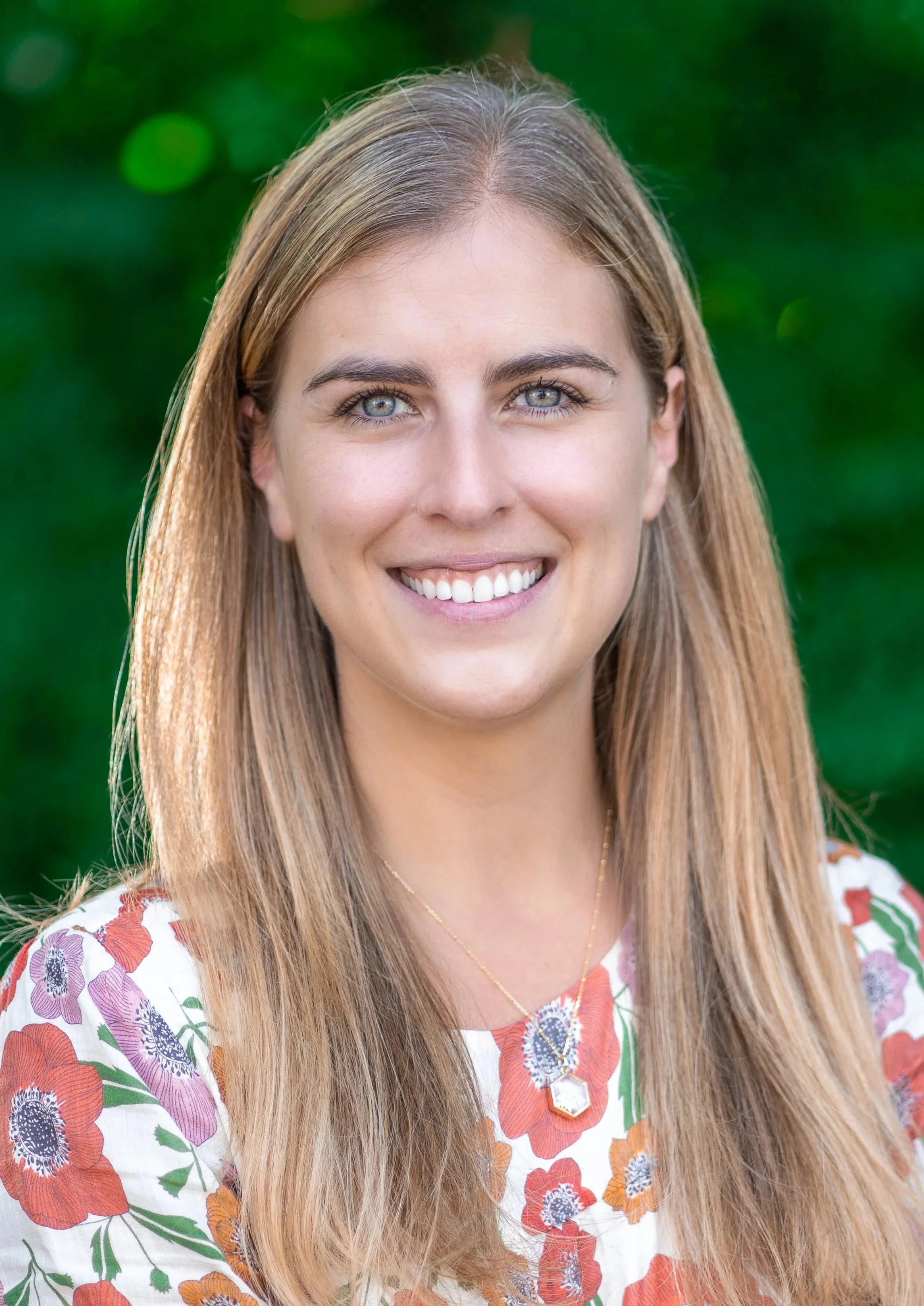A smiling woman with long, light brown hair and blue eyes outdoors, wearing a floral top and a gold necklace.