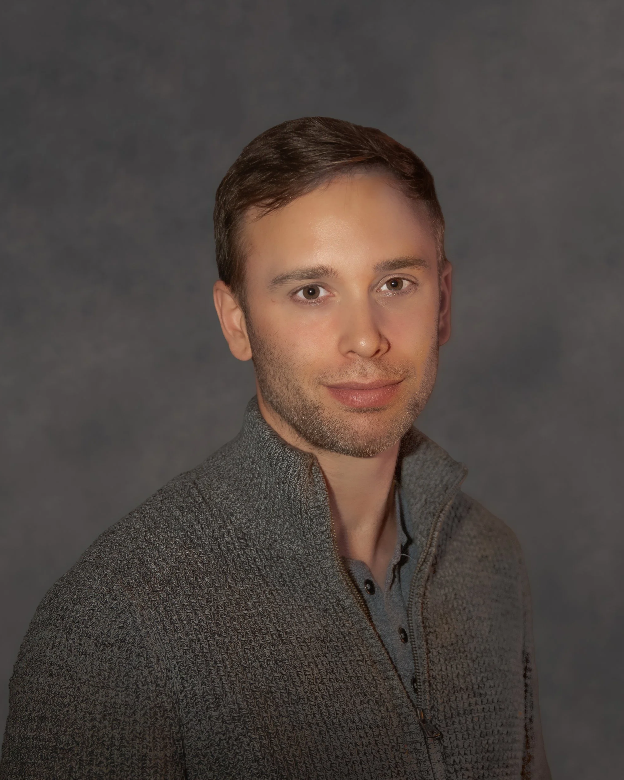 A young man with short brown hair and light skin, wearing a gray, textured zip-up sweater, poses against a dark gray background.