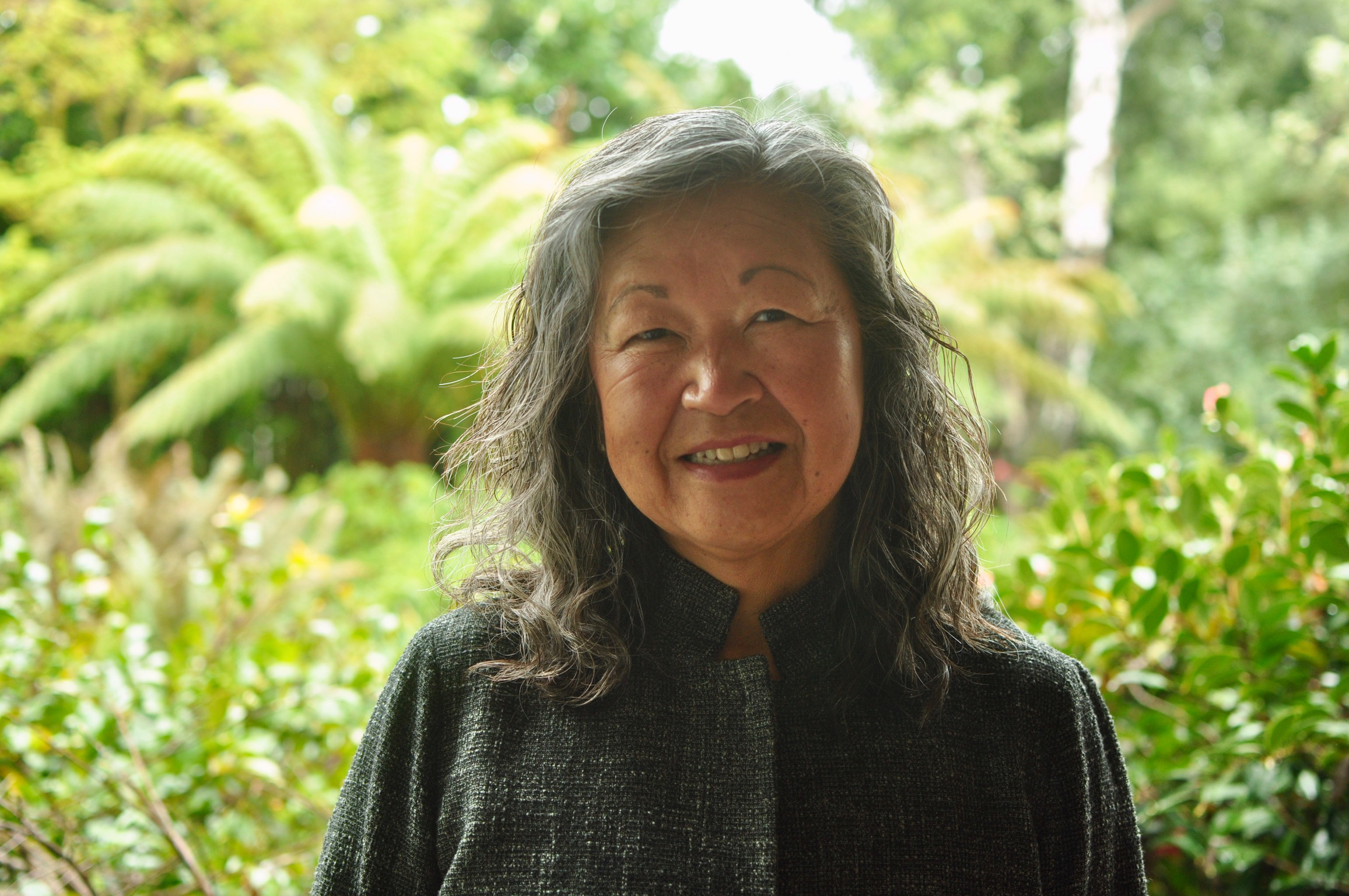 A smiling elderly Asian woman with wavy gray hair standing outdoors surrounded by green plants and trees.