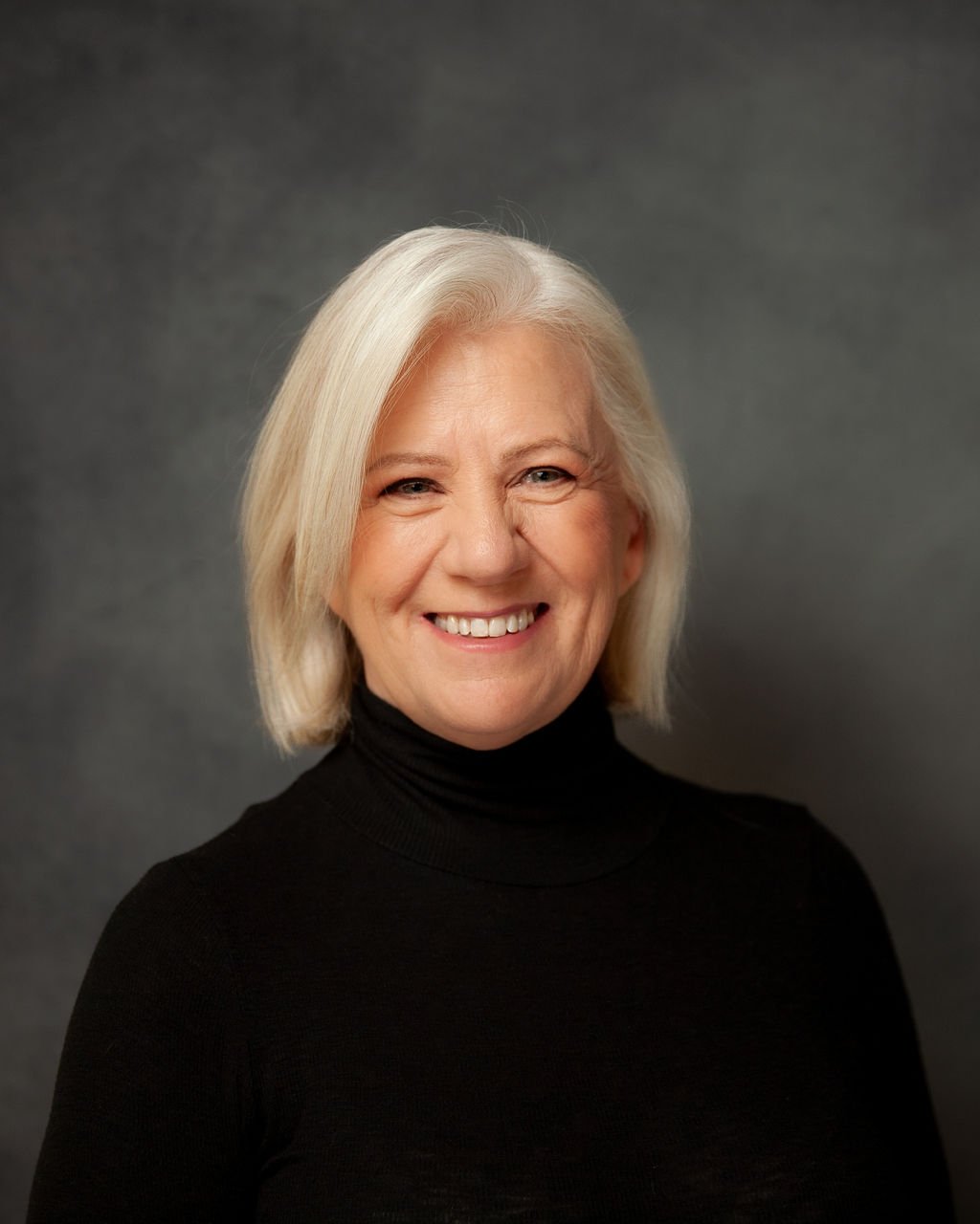 A smiling middle-aged woman with shoulder-length blond hair, wearing a black turtleneck, posing against a dark grey background.