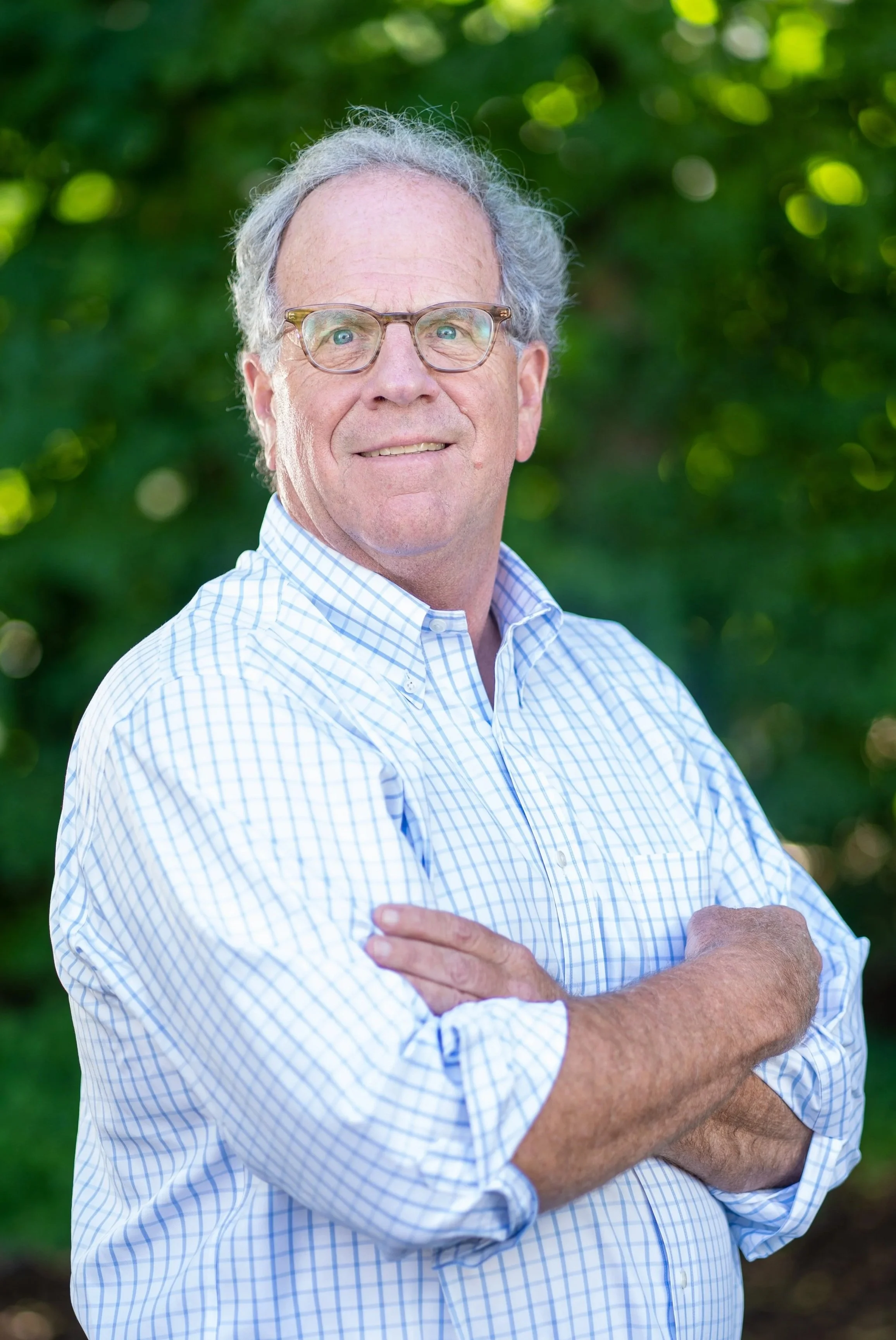 A middle-aged man with gray hair, glasses, and light skin standing outdoors with arms crossed, wearing a blue and white checkered shirt, and smiling. Green leafy background.