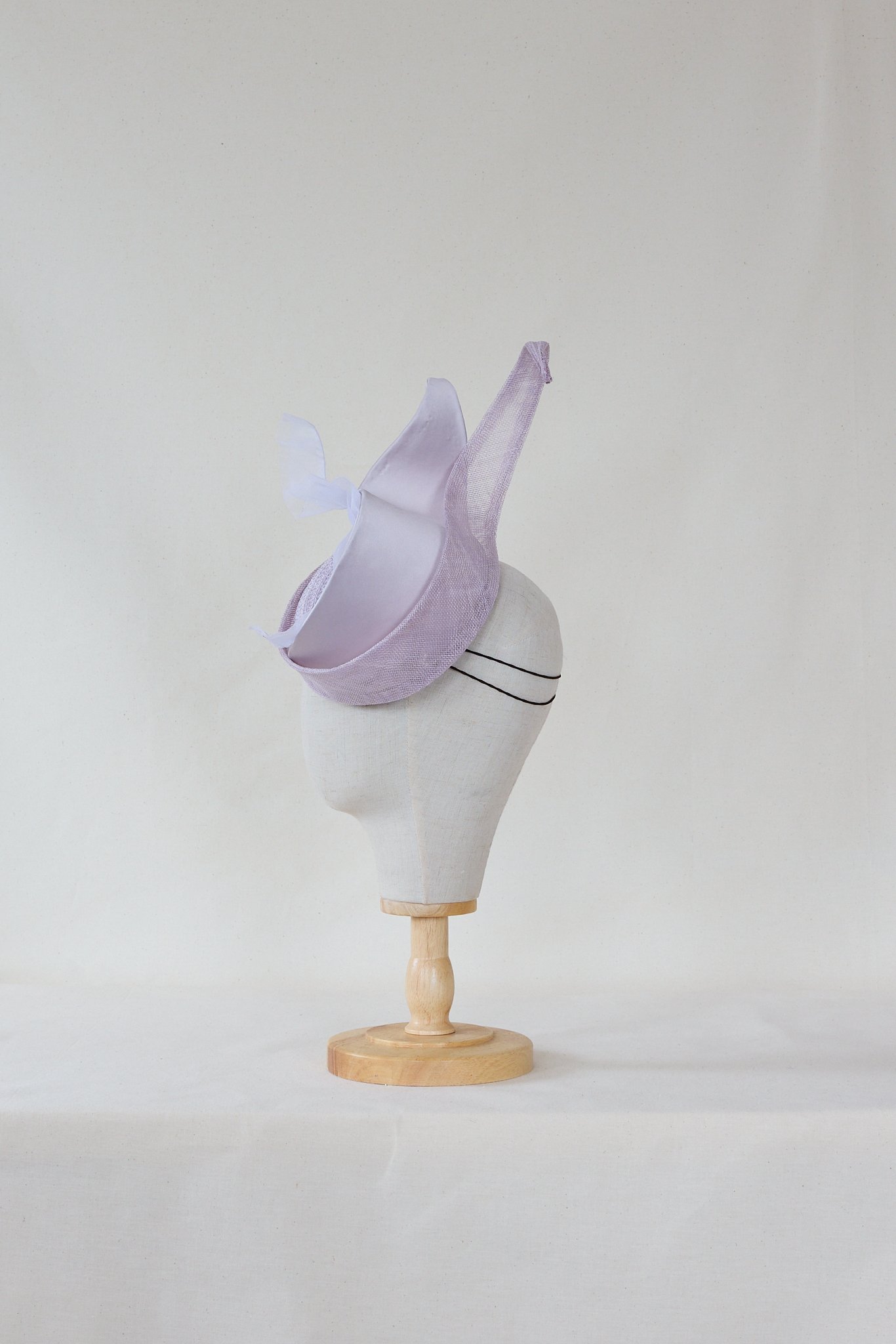 A mannequin head wearing a light purple fascinator hat with a large bow and netting, mounted on a wooden stand against a plain white background.