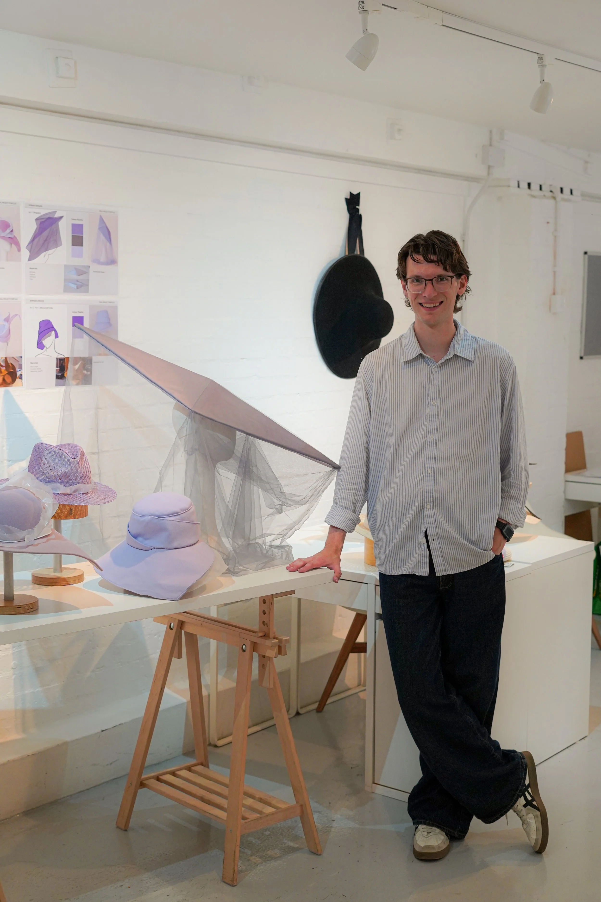 A young man with glasses, in a light striped shirt and dark pants, smiling and leaning on a white table with hats and fabric displayed, in a bright art or design studio.