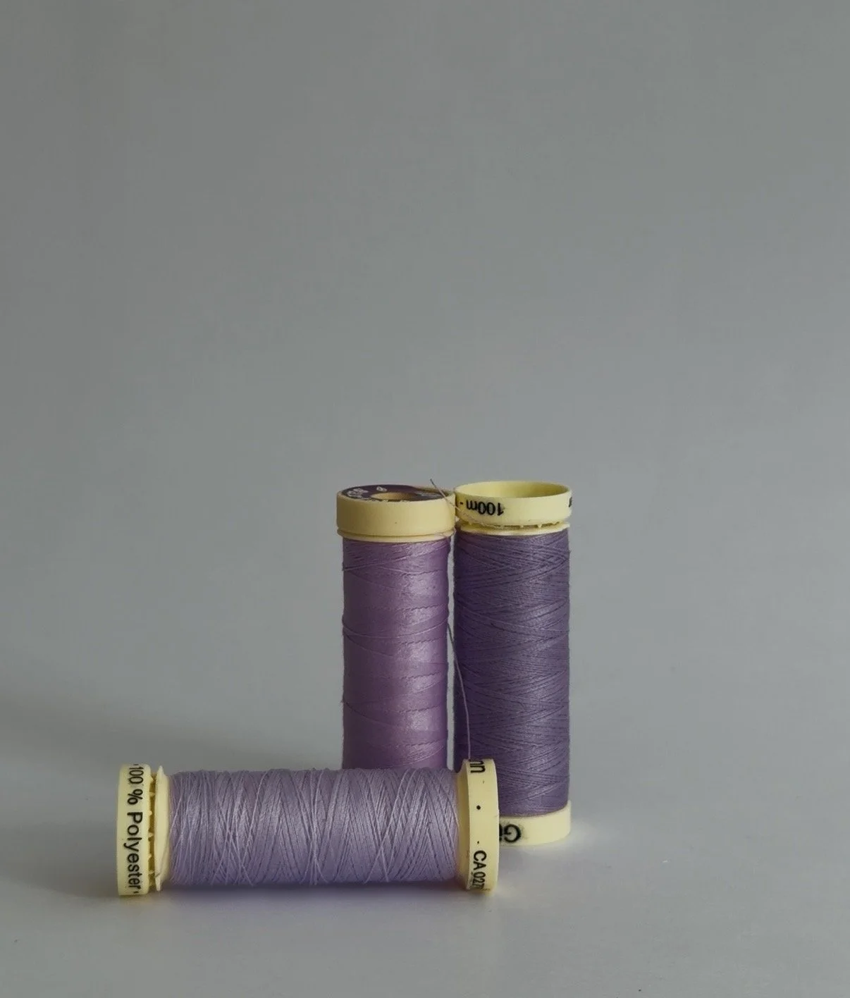 Three spools of light purple sewing thread arranged against a gray background, with one spool lying horizontally and two standing vertically.