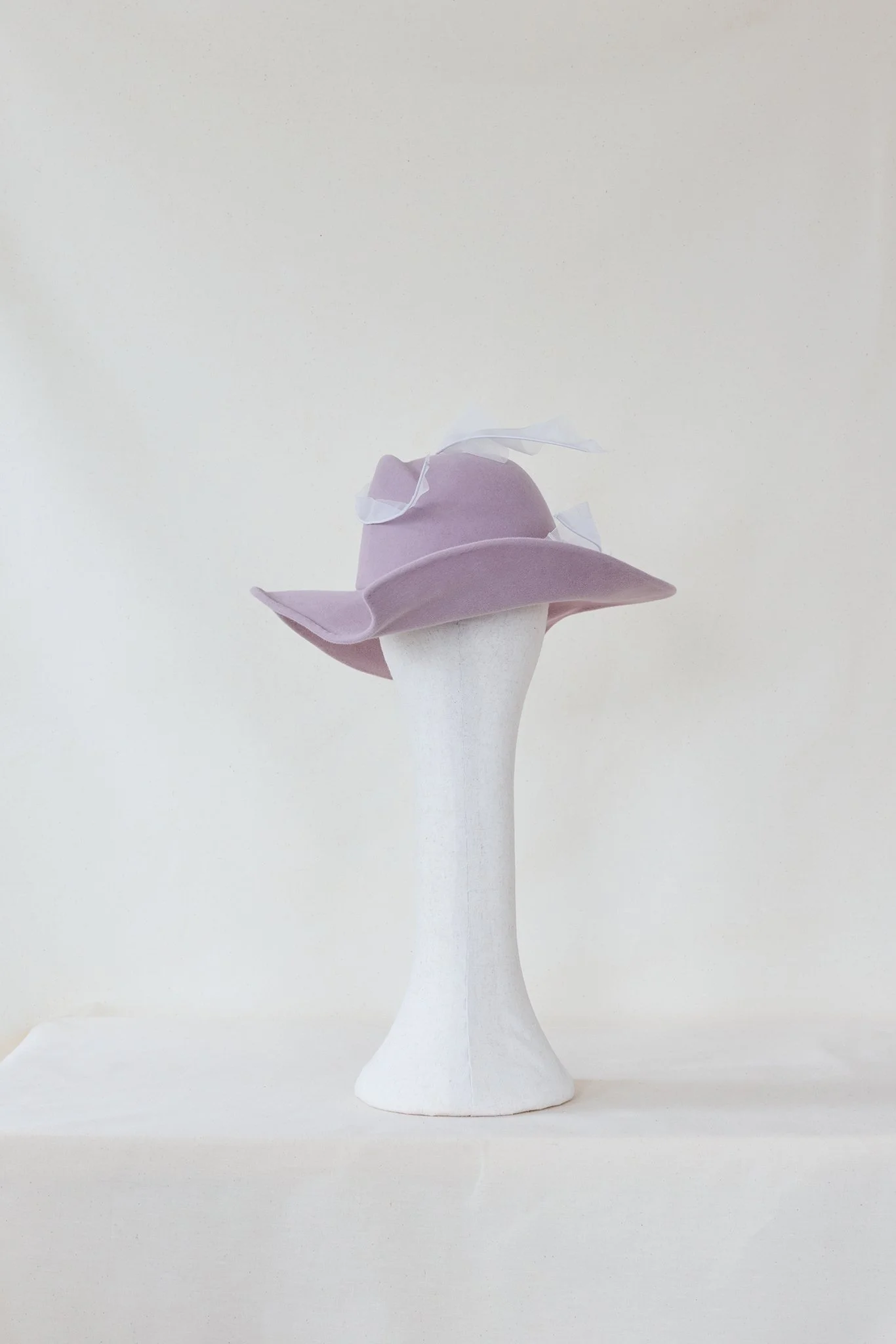 A mannequin head wearing a lavender wide-brimmed hat with a white ribbon, set against a plain, neutral background.