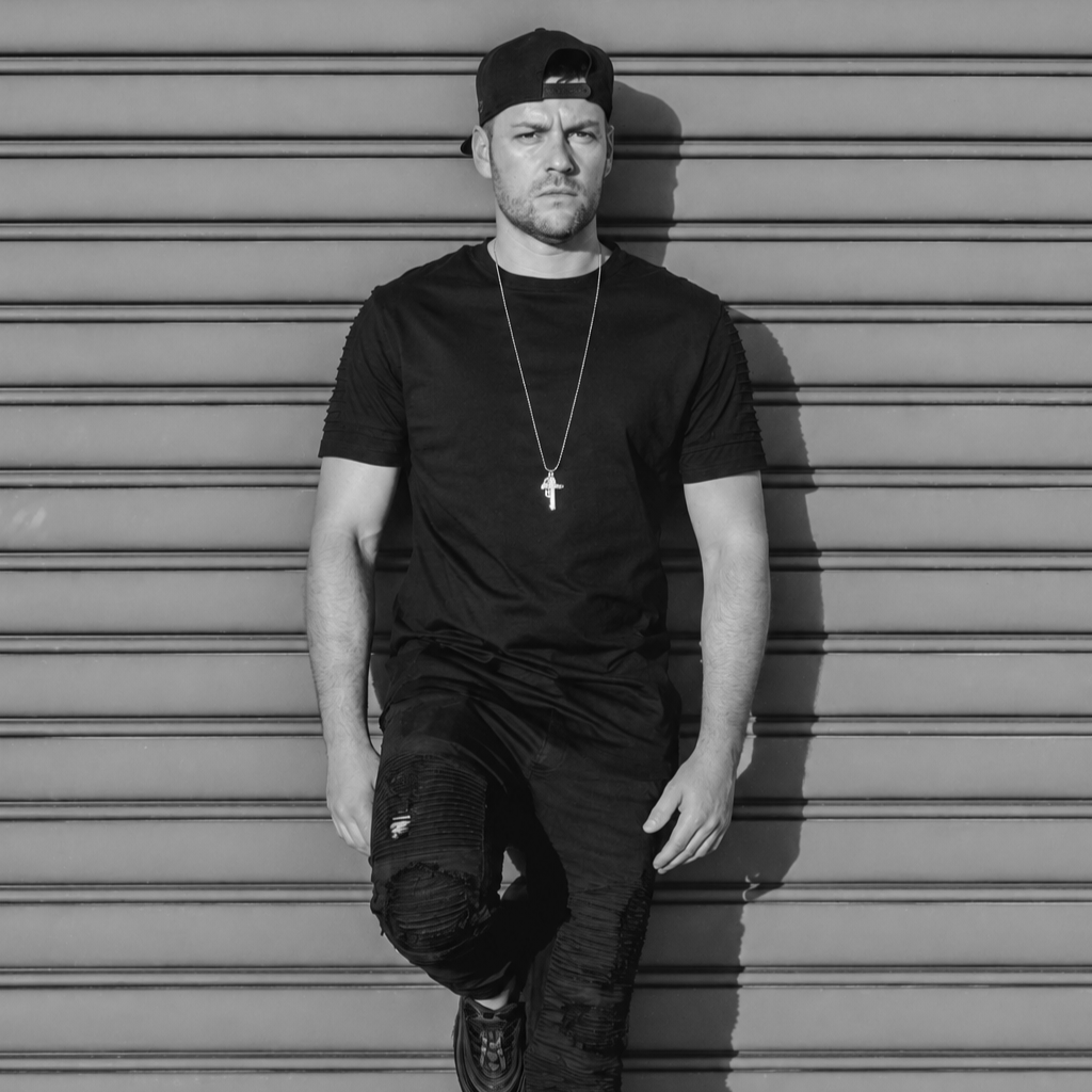 A young man with a serious expression, wearing a black t-shirt, ripped jeans, a backwards black cap, a necklace with a cross pendant, and sneakers, standing against a metal shutter in black and white.
