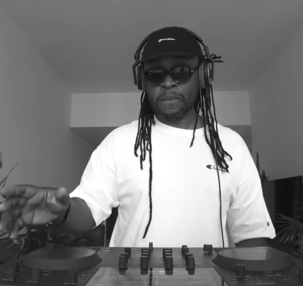 A man with dreadlocks wearing sunglasses, a cap, and a white t-shirt is DJing with a turntable and mixer in front of him, in a room with plain walls.