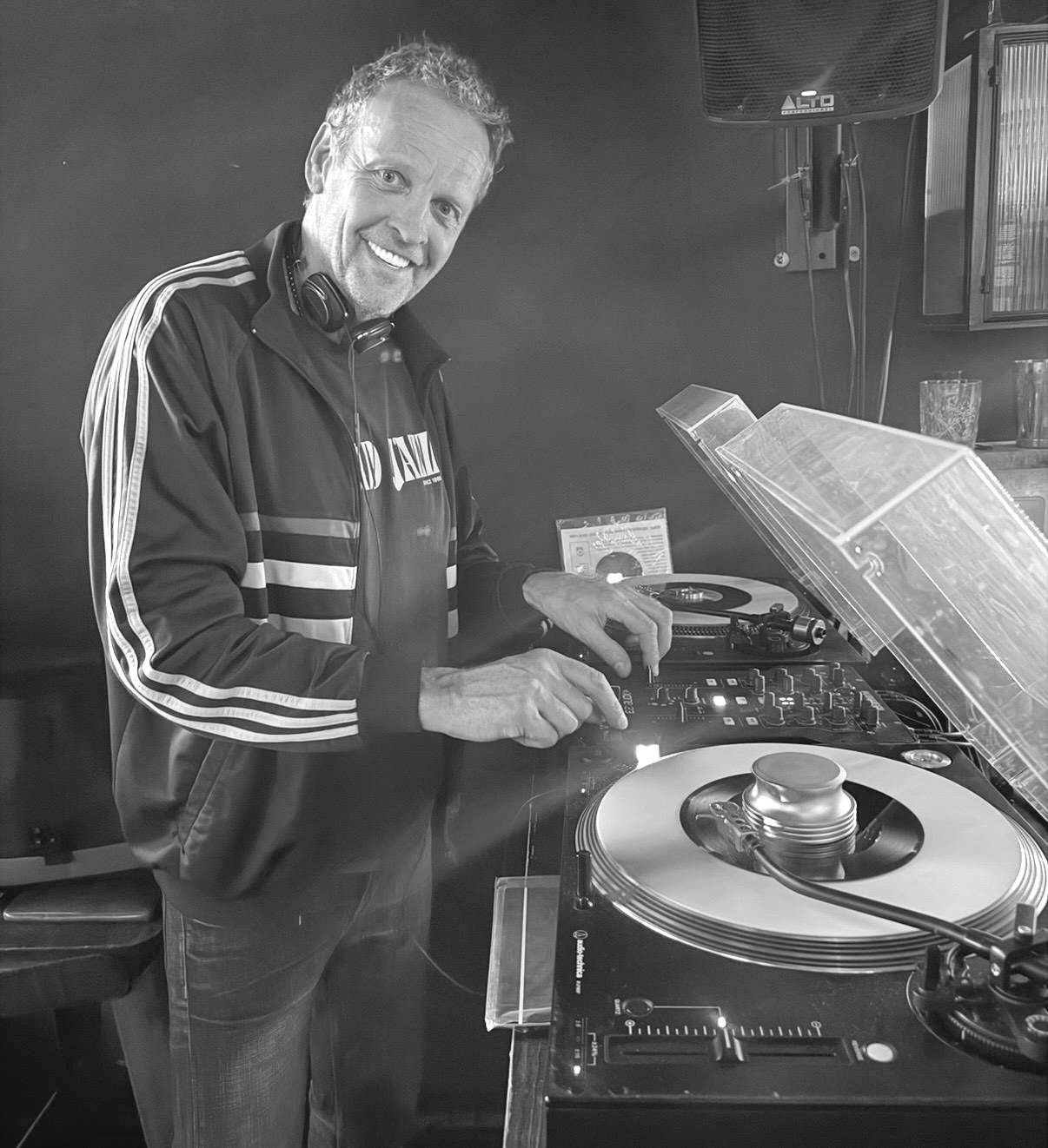 A man with headphones around his neck is DJing at a turntable setup, smiling at the camera in a black and white photo.
