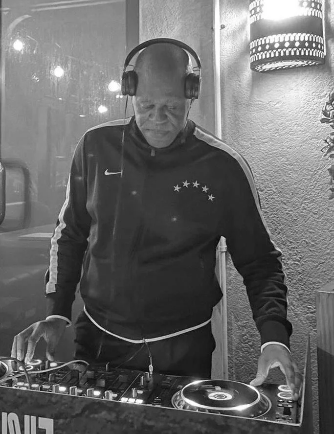 A man wearing headphones and a dark sports jacket with stars on the chest, standing behind DJ equipment in a dimly lit room.