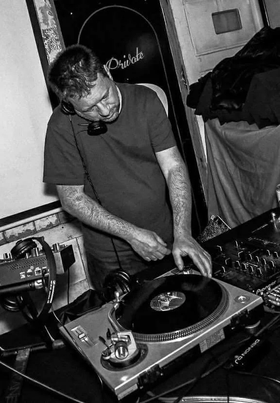 A man wearing a t-shirt and headphones, with curly hair, is mixing music on a DJ turntable setup in a dimly lit room.