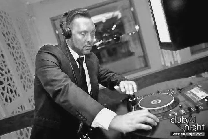 A man in a suit and tie DJing at a nightclub, wearing headphones and working on a DJ mixing console.