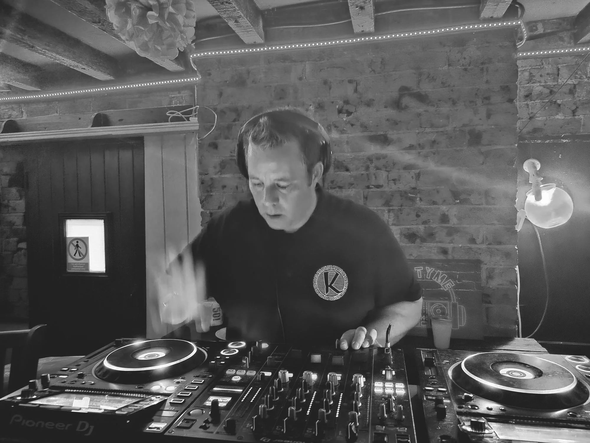 A DJ is mixing music at a nightclub, wearing headphones and standing behind a DJ mixer with turntables. The setting is indoors with a brick wall background, and there is a small wall lamp on the right.