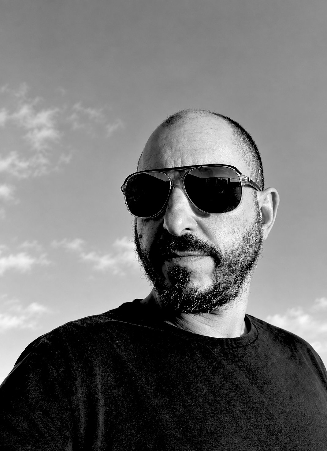 A man with a beard and shaved head wearing dark sunglasses and a black t-shirt outdoors with a partly cloudy sky in the background.