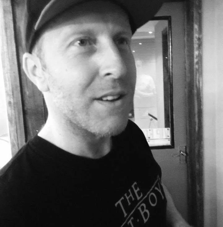A man with light skin, wearing a black T-shirt and a cap, standing indoors near a door with a window.