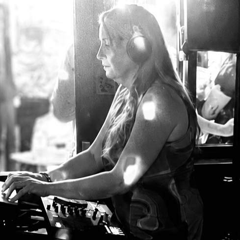 A woman with long hair wearing headphones, operating DJ equipment in a dimly lit setting.