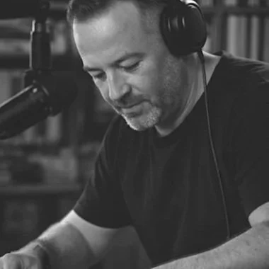 A man wearing headphones, sitting at a desk, speaking into a microphone, with bookshelves in the background.
