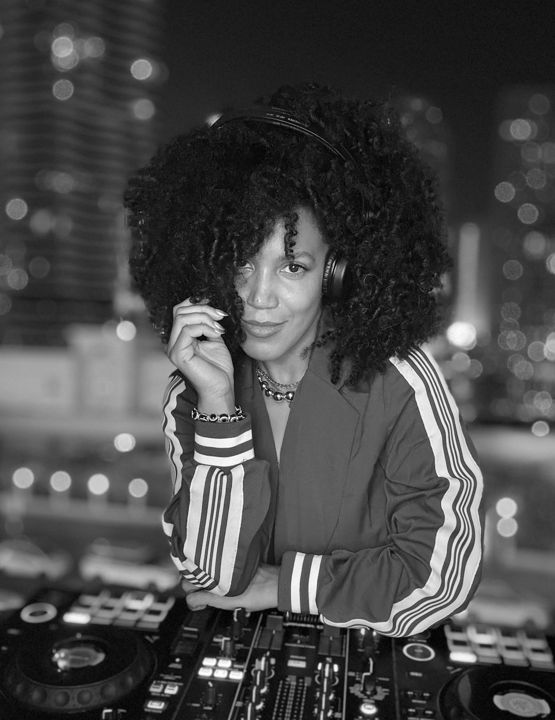 A woman with curly hair wearing headphones behind DJ equipment, with city lights in the background.