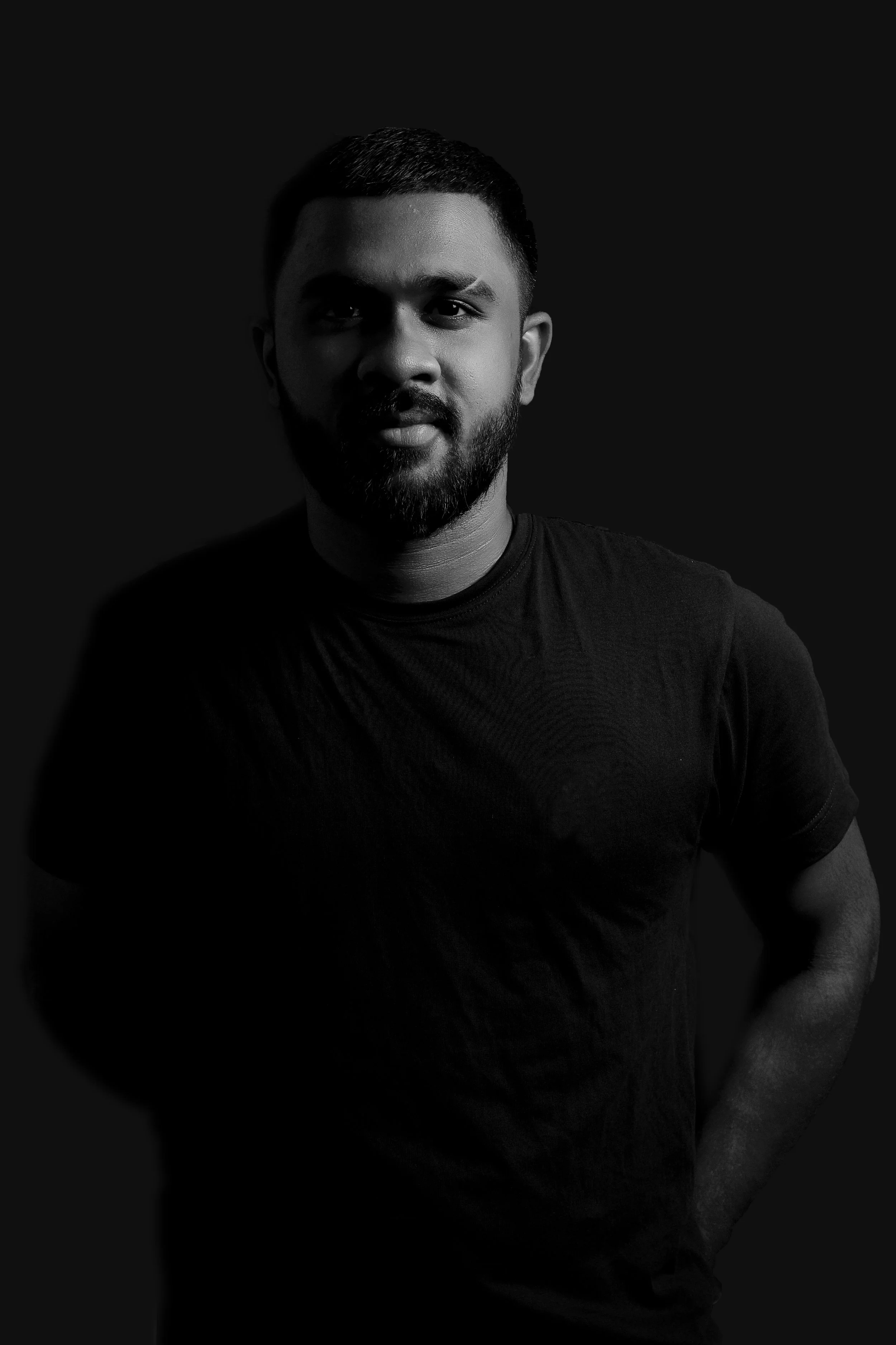 A man with a beard and short hair wearing a black t-shirt, standing against a dark background.