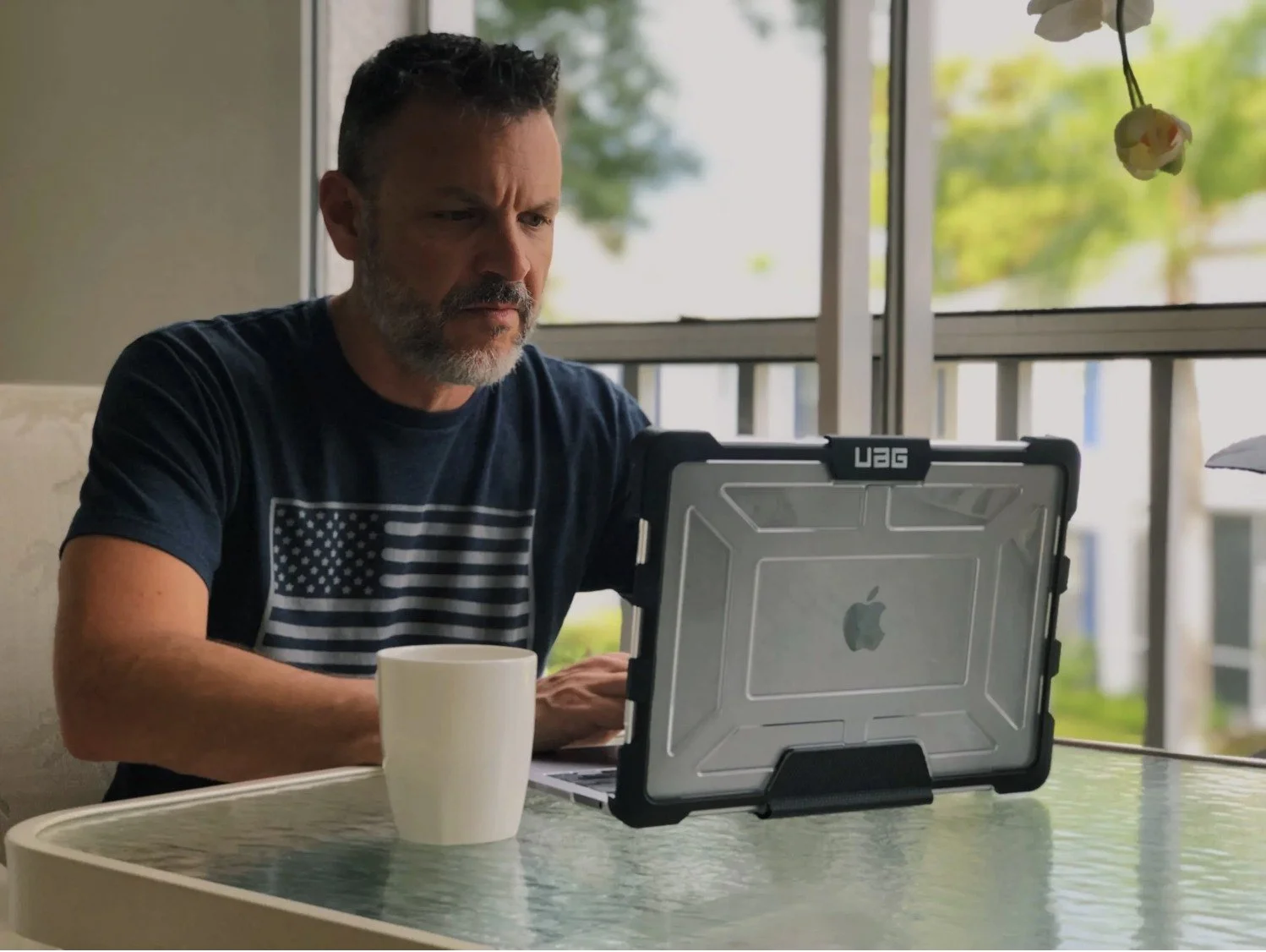 A man with a beard sitting at a table, looking at a laptop with an Apple logo, next to a white mug, in a sunny room with large windows.