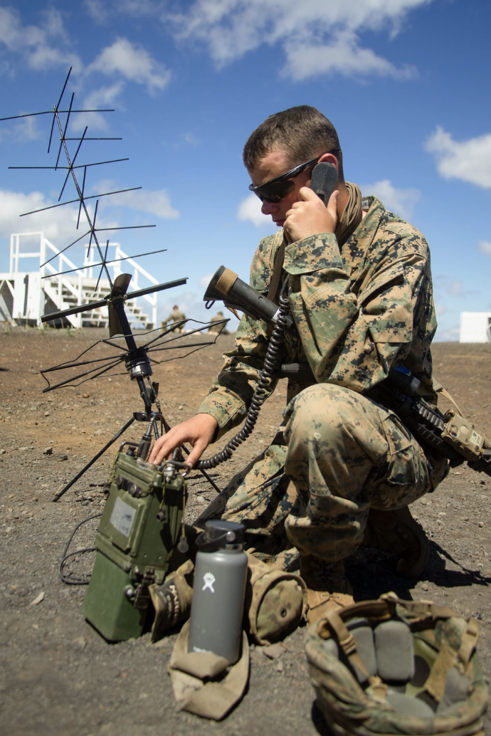 A soldier in camouflage uniform kneeling on the ground, wearing sunglasses, talking on a radio handset, with radio equipment and antennas nearby, outdoors on a clear day.