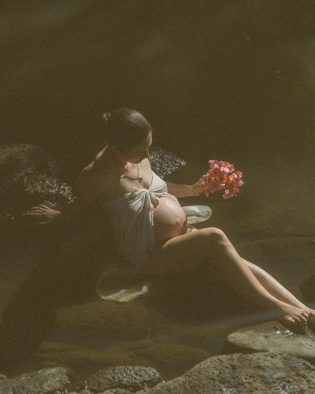 A pregnant woman sitting on rocks in the water, holding a bouquet of pink flowers, with her head slightly bowed down.