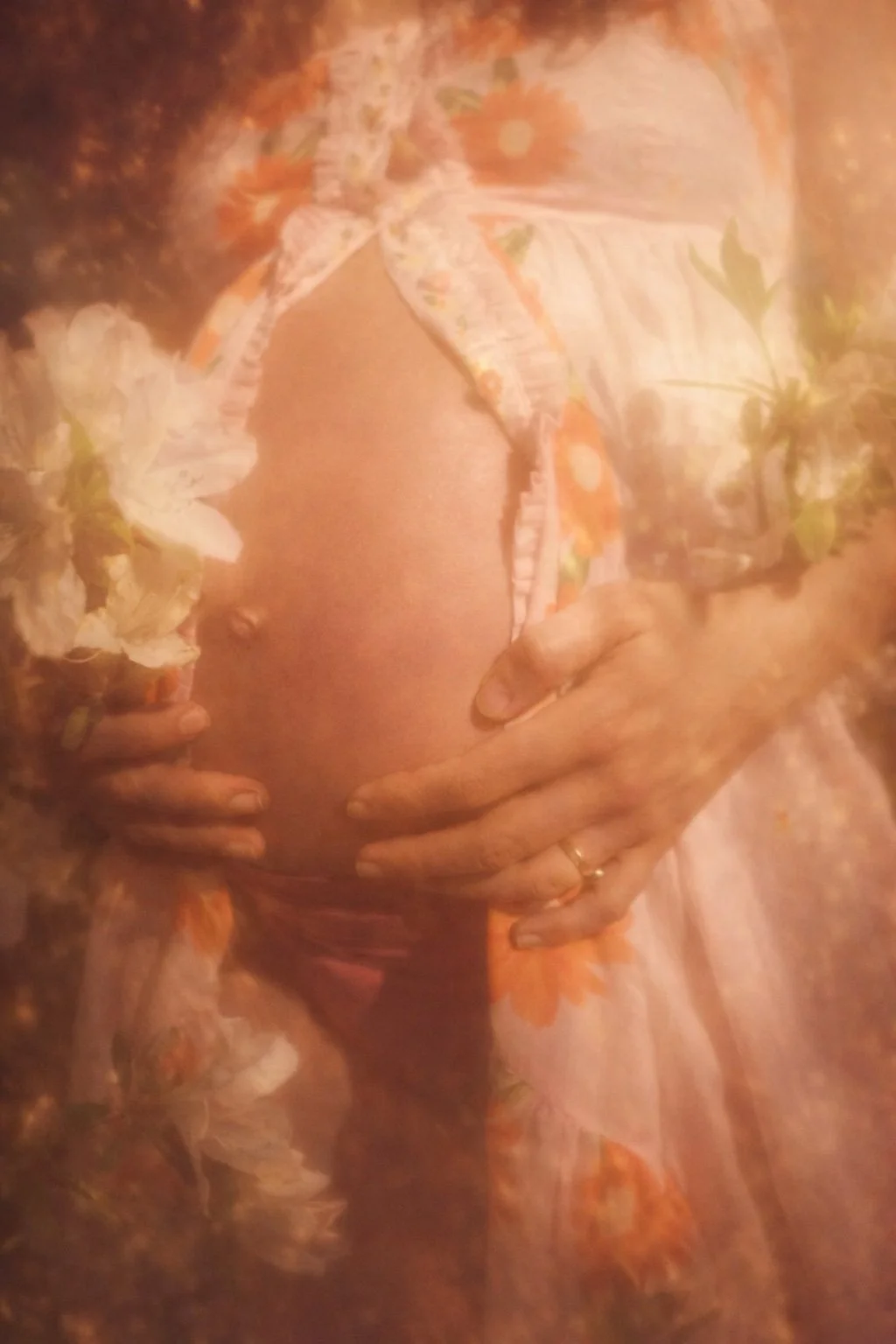 A pregnant woman wearing a floral dress, with her hands resting on her belly, in a softly lit setting.