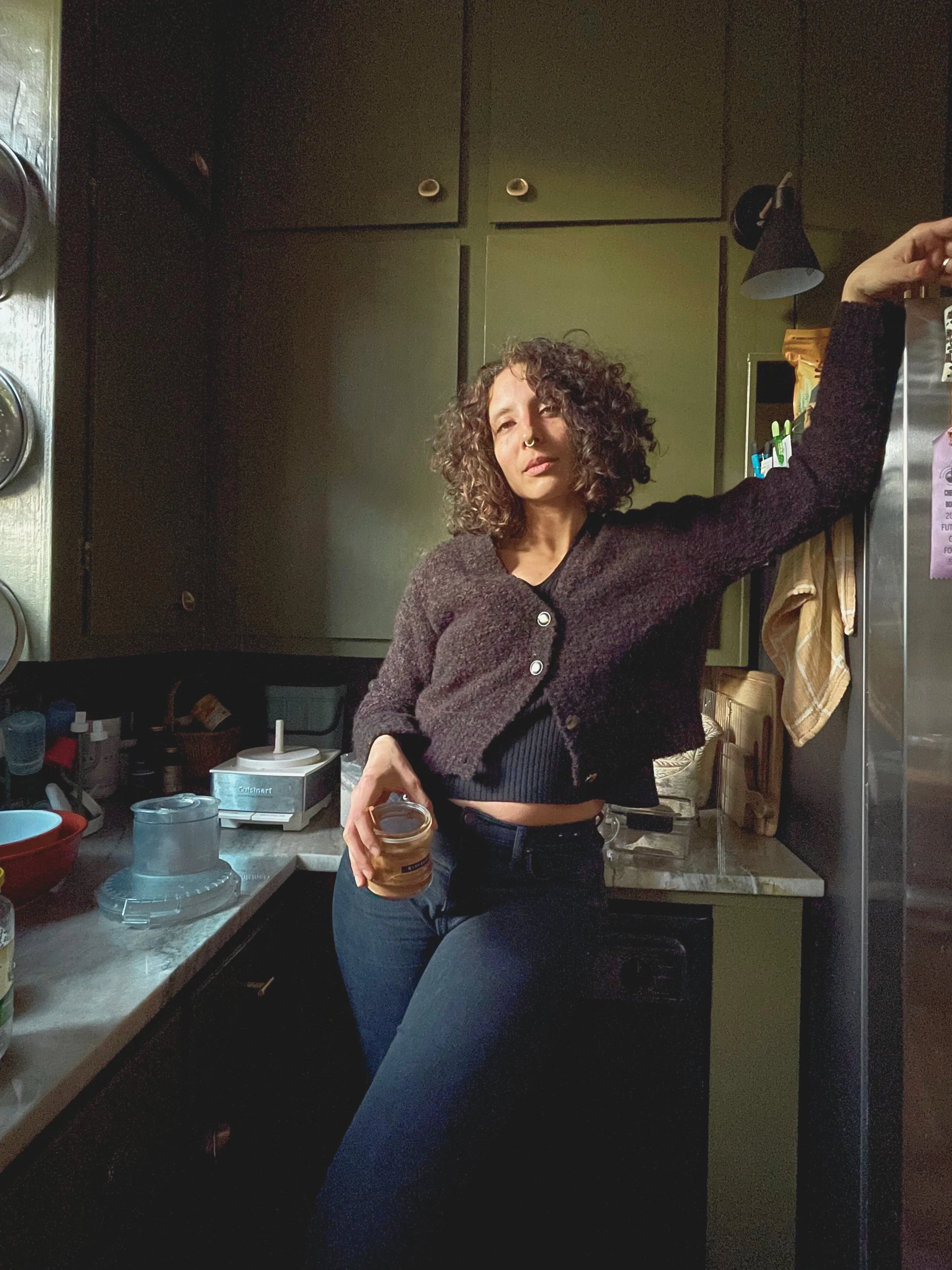 A woman with curly hair, wearing a dark cardigan and jeans, standing in a kitchen, holding a jar and leaning against a refrigerator.