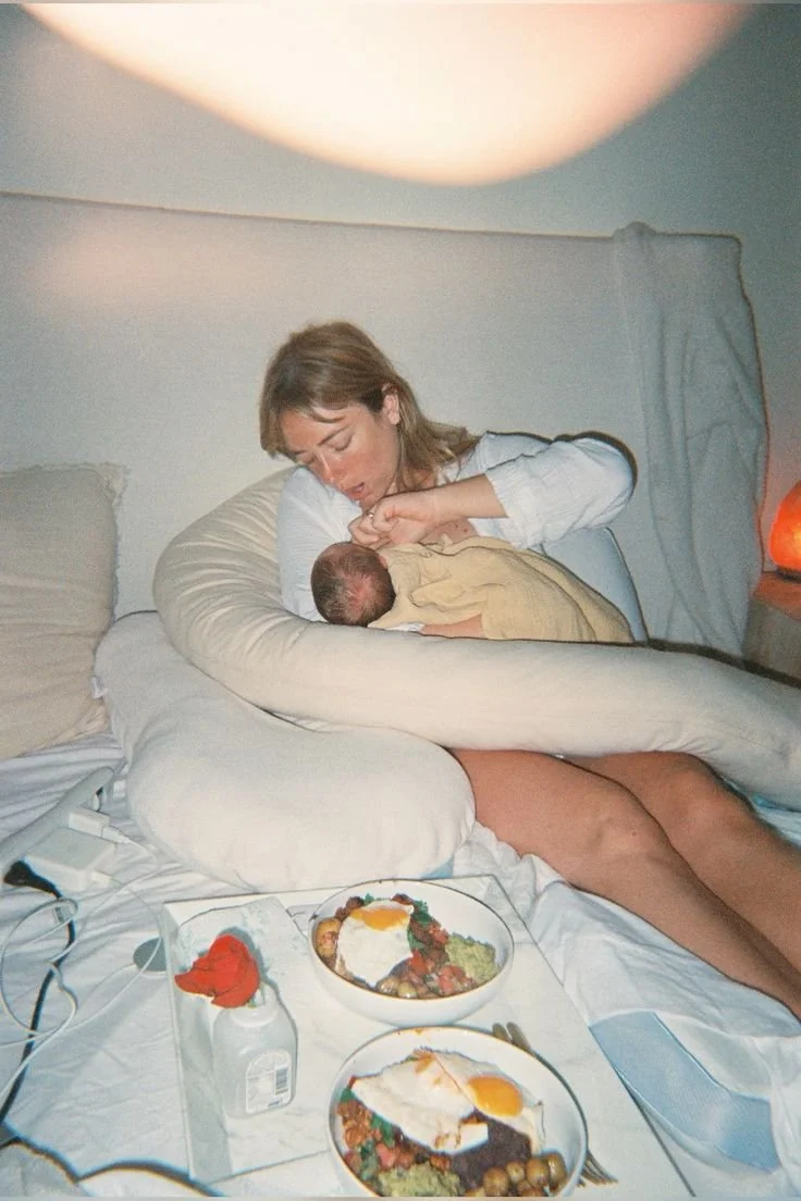 Woman breastfeeding a newborn in a cozy bedroom with plates of breakfast on the bed.