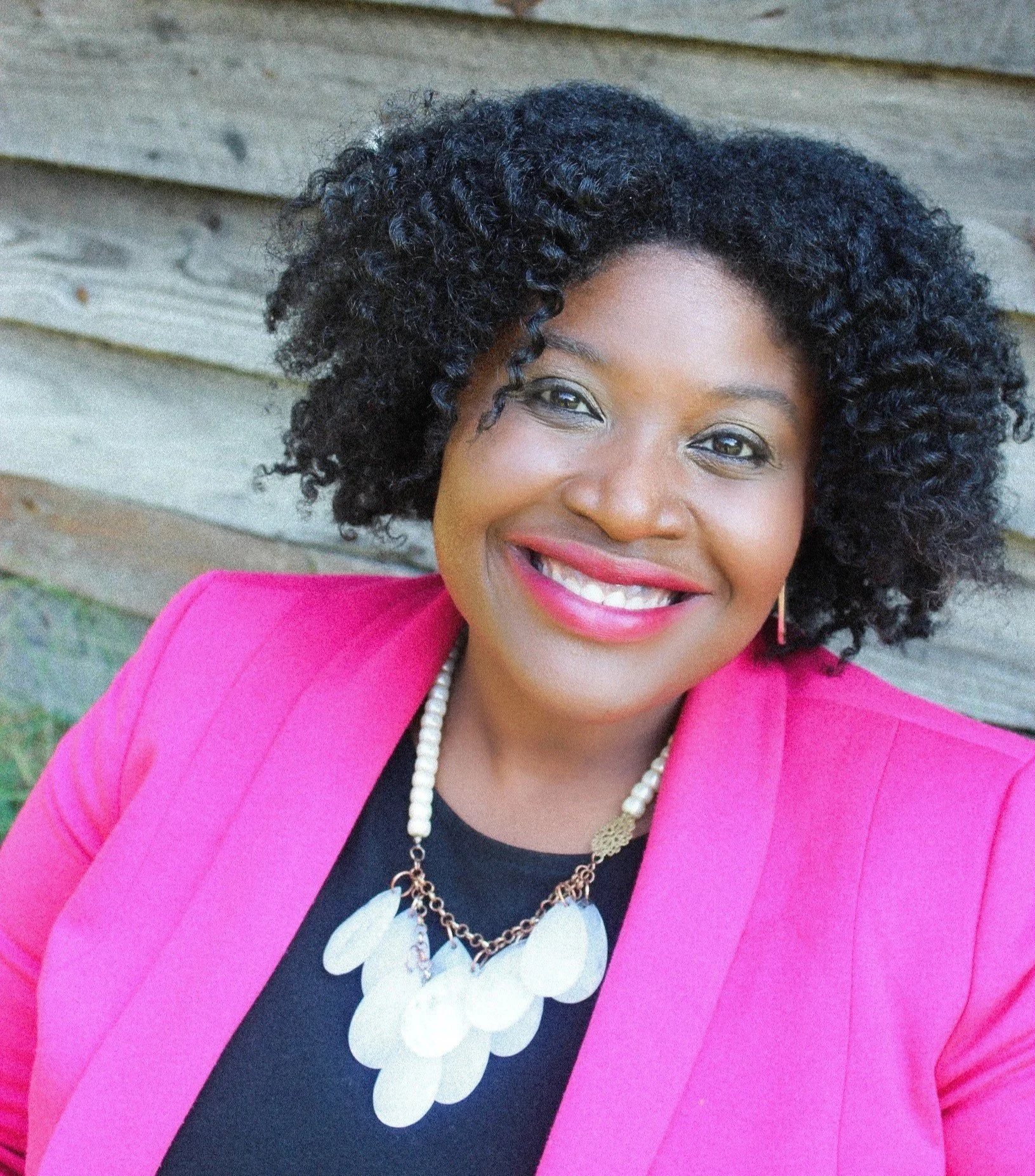 A smiling woman with curly black hair, wearing a pink blazer, black top, white pearl necklace, and gold earrings, standing outdoors in front of a wooden fence.