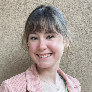 A young woman with grayish-brown hair, wearing a pink jacket, smiling in front of a beige textured wall.